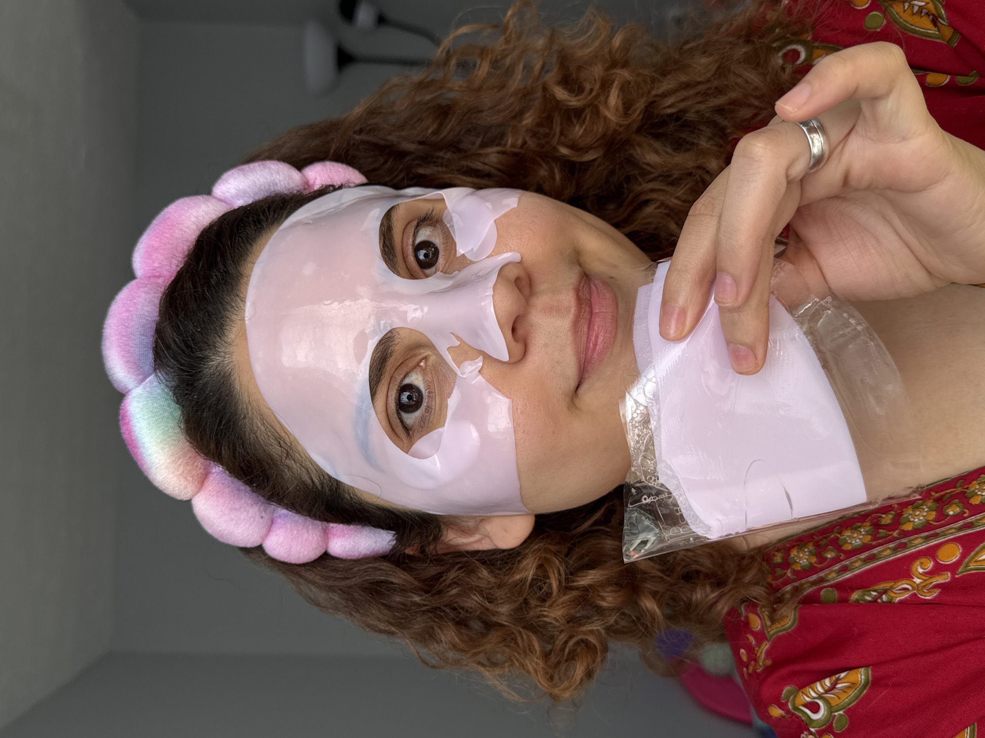 The image shows a person with curly brown hair wearing a pink and multi-colored headband and a white sheet mask on their face. They are holding the mask packaging in their hand, which has a silver ring on one of the fingers. The person is wearing a red top with a yellow and green pattern. The background appears to be a room with a gray wall. There is no information to suggest this photo was taken in Columbus, United States of America.