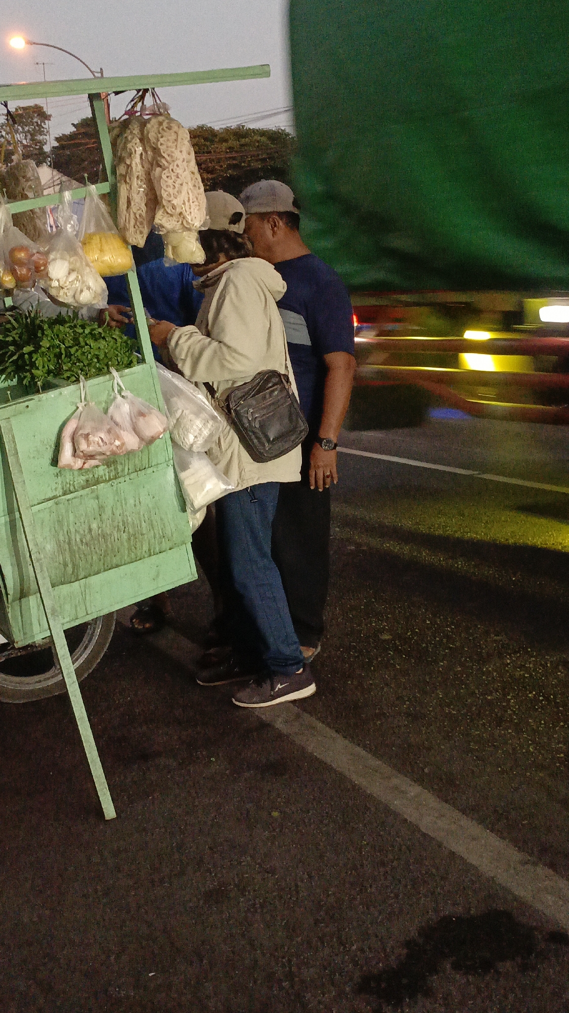 In Srono, Indonesia, three individuals stand near a mobile food cart at dusk. The cart, painted green, displays various food items in plastic bags, including what appear to be vegetables, meat, and dried goods. A person in a light-colored jacket and blue jeans is interacting with the cart, holding plastic bags. To their right, a person in a dark blue shirt and a baseball cap leans in, their arm around the other person. A third person, partially obscured, stands behind the cart. The background shows a road with blurred vehicle lights and a large green tarp. The overall lighting suggests it is either early morning or late evening.
