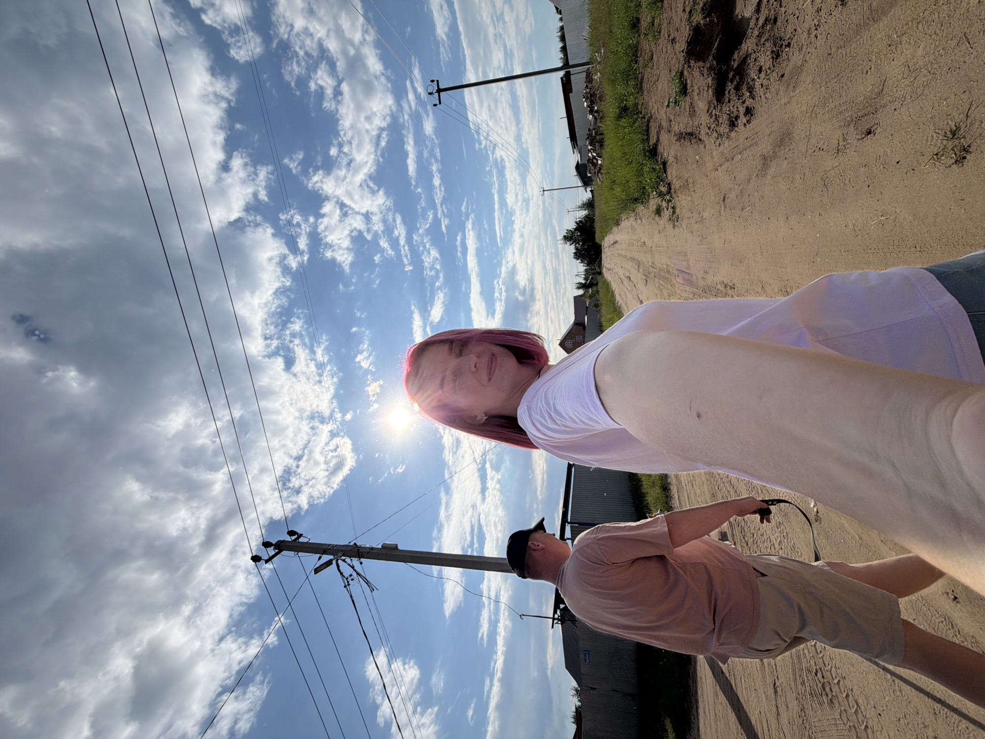 In Chaikovsky, Russia, on a dirt road lined with green grass and distant houses, a woman with short, pink hair is taking a selfie. She is wearing a white t-shirt and is partially visible in the foreground. Behind her, a man in a beige t-shirt and tan shorts is walking away from the camera, holding a black leash. The sun is bright, casting lens flare into the sky, and there are scattered white clouds against a blue sky. Overhead, power lines are strung between utility poles. The overall scene depicts a casual outdoor moment on a sunny day.