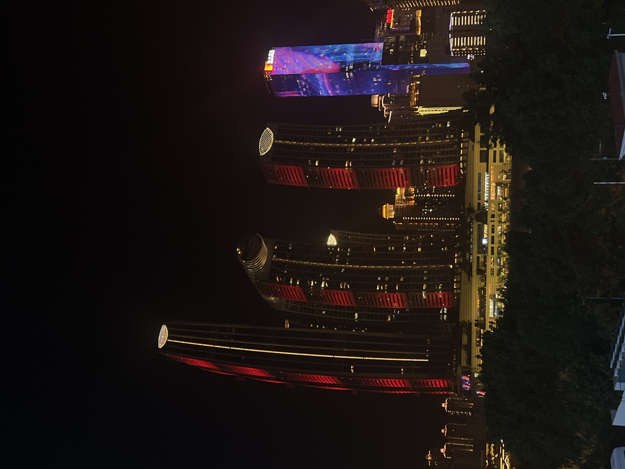 The image depicts a nighttime cityscape with multiple illuminated skyscrapers in Jiangbeicheng Subdistrict, China. The foreground features dark foliage, likely trees, obscuring the lower portions of some structures. Dominating the scene are several tall buildings, some featuring red and yellow vertical lighting accents, while others display vibrant blue and pink patterns. One particularly striking skyscraper on the right is adorned with a facade that glows with a dynamic mix of blue and pink lights, featuring vertical stripes and abstract patterns. To its left, a taller building is primarily illuminated with red horizontal bands and a glowing circular element at its apex. Several other buildings of varying heights are visible in the background, all lit up, contributing to a bustling urban night atmosphere. The sky is a deep, dark black, typical of a nighttime setting. No people or specific actions are discernible due to the distance and the focus on the architectural illumination.