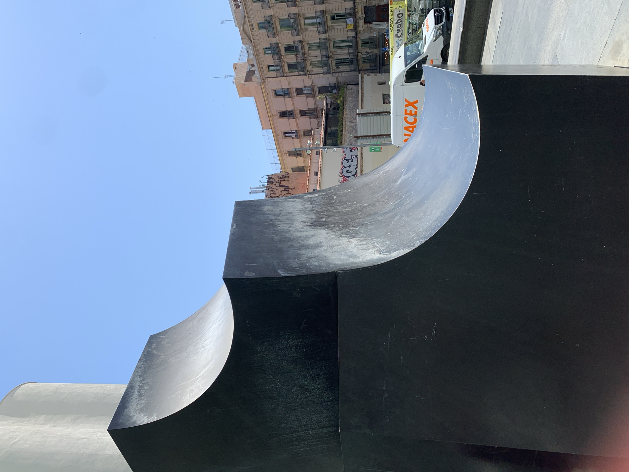 The image captures a bright, sunny day in Barcelona, Spain. In the foreground, a large, dark, angular sculpture with a curved surface dominates the scene. Its surface appears to be made of metal, reflecting the light and showing signs of wear. Behind the sculpture, a white van with "NACEX" branding is visible. Further in the background, a row of buildings with balconies and windows line a street. The sky is clear and blue. The overall impression is an urban outdoor setting on a clear day.