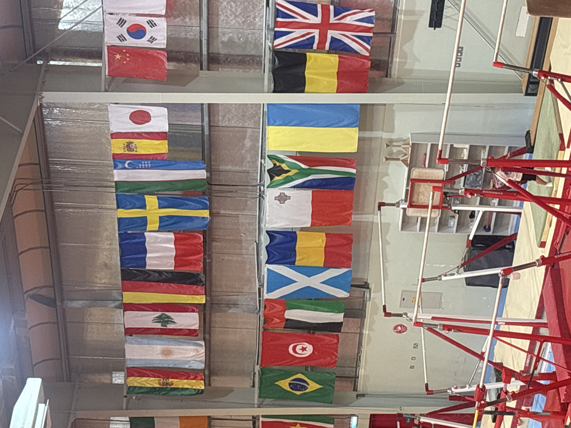 The image depicts an indoor gymnasium, likely a gymnastics facility, with various flags hung from the ceiling. The flags represent different countries and are displayed in two rows. The gymnasium features red and white gymnastics equipment, including uneven bars and balance beams. Shelving units filled with sports equipment are visible against the back wall. The lighting suggests an interior environment. There is no visible indication of the city being Dubai, United Arab Emirates, in the image. A young person, possibly a child, is partially visible in the lower right portion of the frame near the gymnastics equipment.