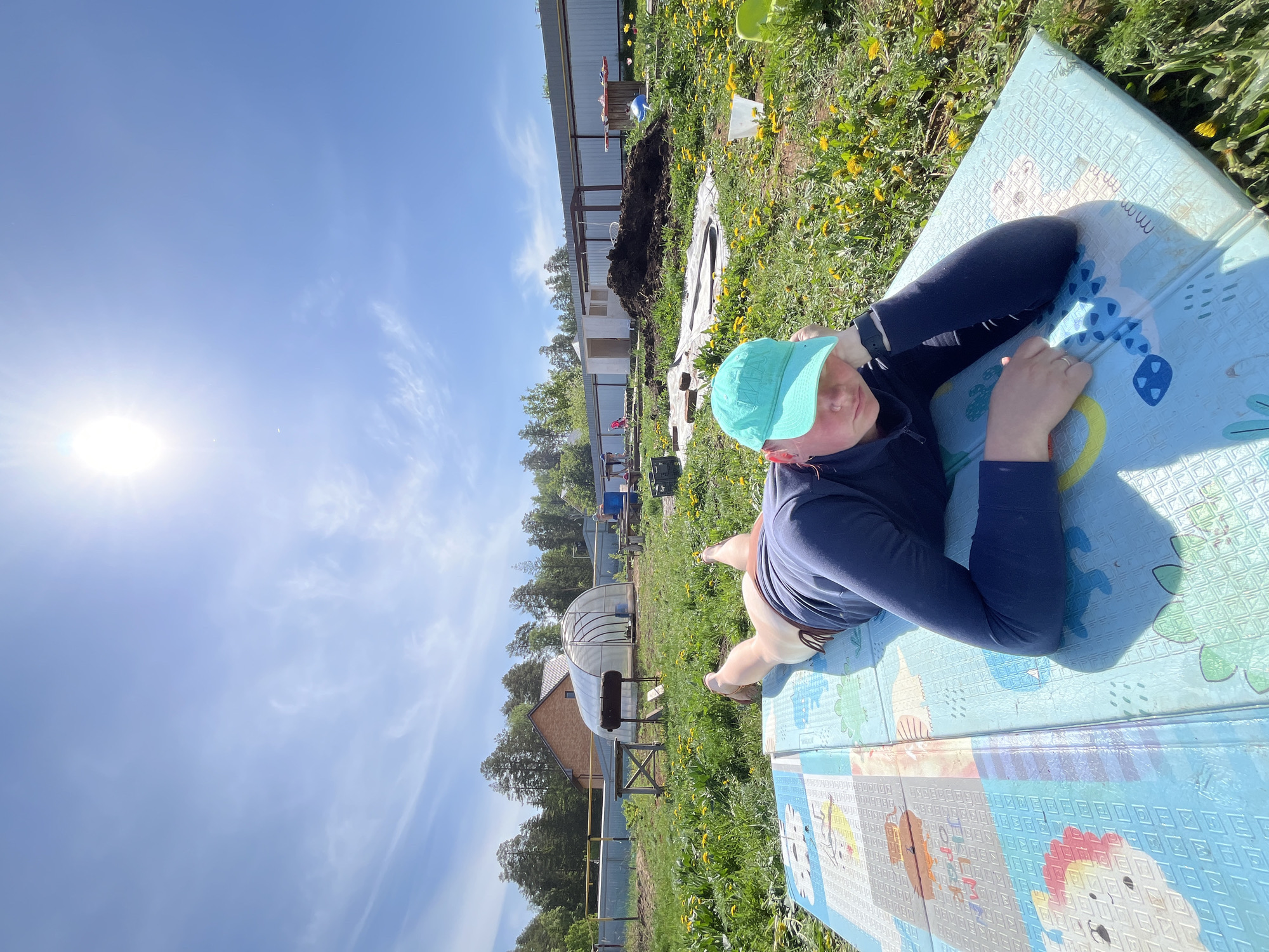 A person is lying on a blue children's play mat in a grassy field dotted with yellow dandelions. The individual is wearing a dark blue long-sleeved shirt and a turquoise baseball cap. The person's legs are visible, clad in light-colored pants. The sky is blue and sunny, with the sun shining brightly on the left side of the frame. In the background, there is a greenhouse, a white-sided building, a pile of dirt covered in white plastic, some trees, and other houses. Based on the visible architecture and general environmental characteristics, the location is consistent with a rural or suburban area, and the details provided are not sufficient to confirm the location in Chaikovsky, Russia. The overall scene portrays a person relaxing outdoors on a sunny day.