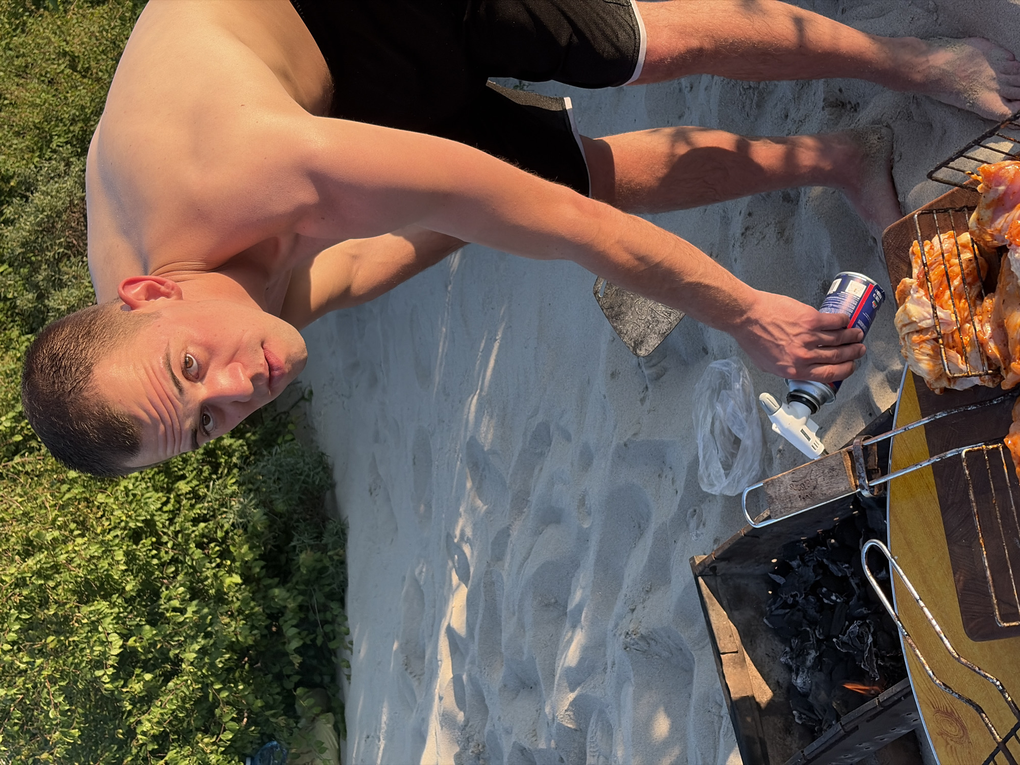 A shirtless man is bent over a grill on a sandy beach. He is holding a can of what appears to be lighter fluid and is spraying it towards the grill, which contains burning coals and raw chicken. The man has short brown hair and is wearing black shorts with white trim. The background consists of dense green foliage and sand. The location is the city of Povorovo, Russia.