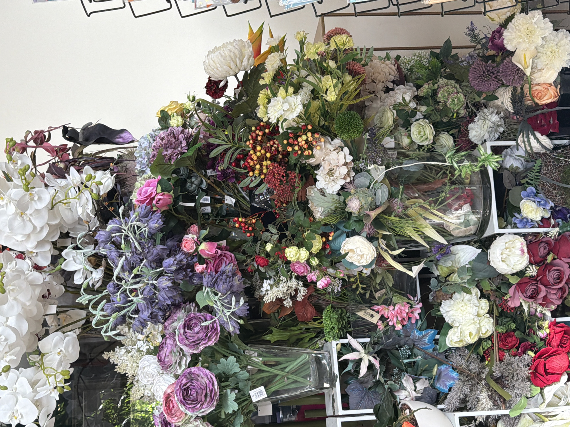 The image shows a display of artificial flowers. The flowers are arranged in bouquets and vases, with a variety of colors and types, including white orchids, purple hydrangeas, pink roses, and red berries. The display appears to be set up in a store or market, with shelves and racks visible in the background. The lighting is bright, and the overall impression is one of abundance and color. The scene is static, with no people or actions visible. The location is unknown, as specified by the prompt.