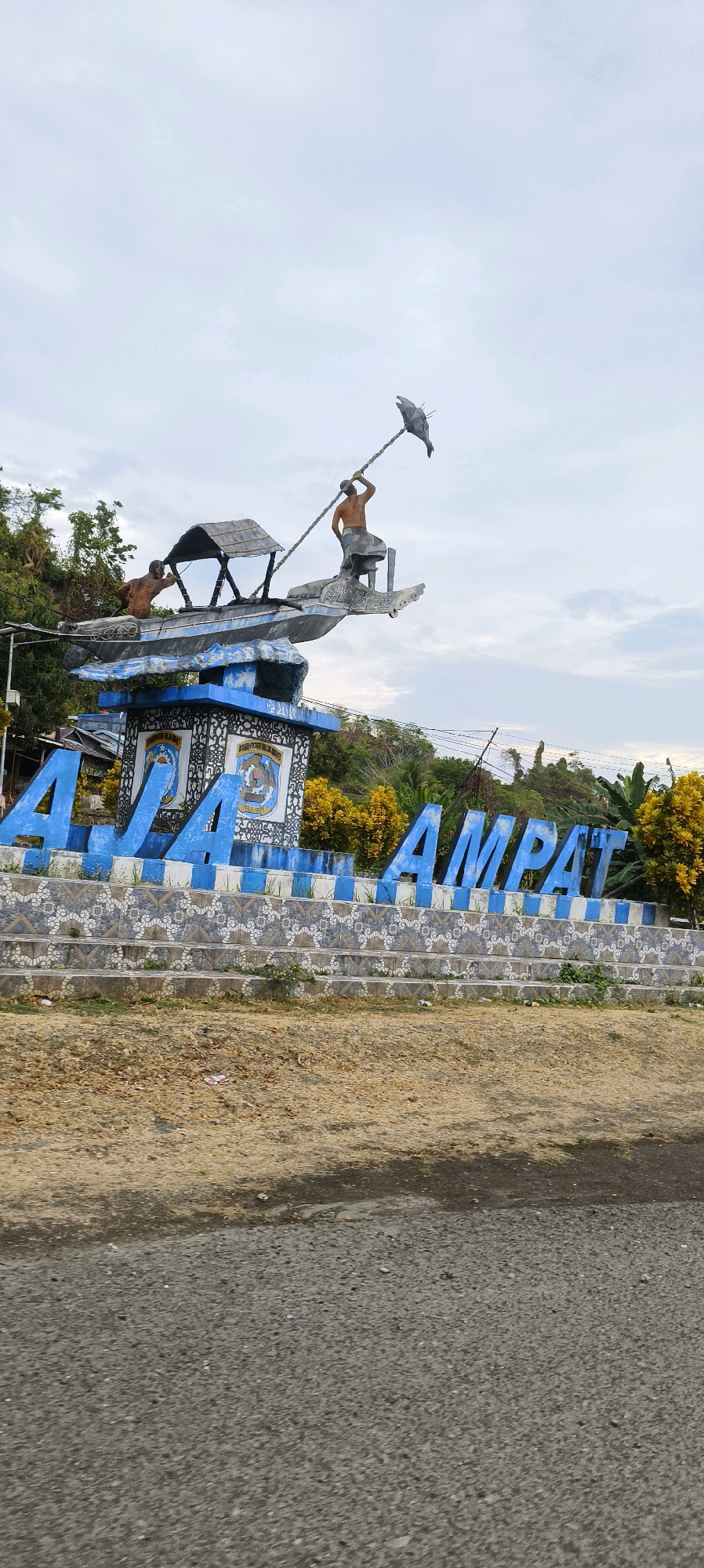 The image displays a monument featuring the words "Raja Ampat" in large blue letters. The monument depicts a boat with two figures, presumably fishermen, engaged in an activity that appears to be related to fishing or hunting, possibly with a bird statue. One figure is in the process of casting a net or spear towards the bird statue, while the other is positioned within the boat. The monument is situated in an outdoor environment with a cloudy sky and lush green foliage in the background. The base of the monument is decorated with intricate blue and white patterns. The foreground consists of dry, rocky ground and a paved road. The monument is a prominent landmark in Waisai, Indonesia, a region known for its natural beauty and marine biodiversity.