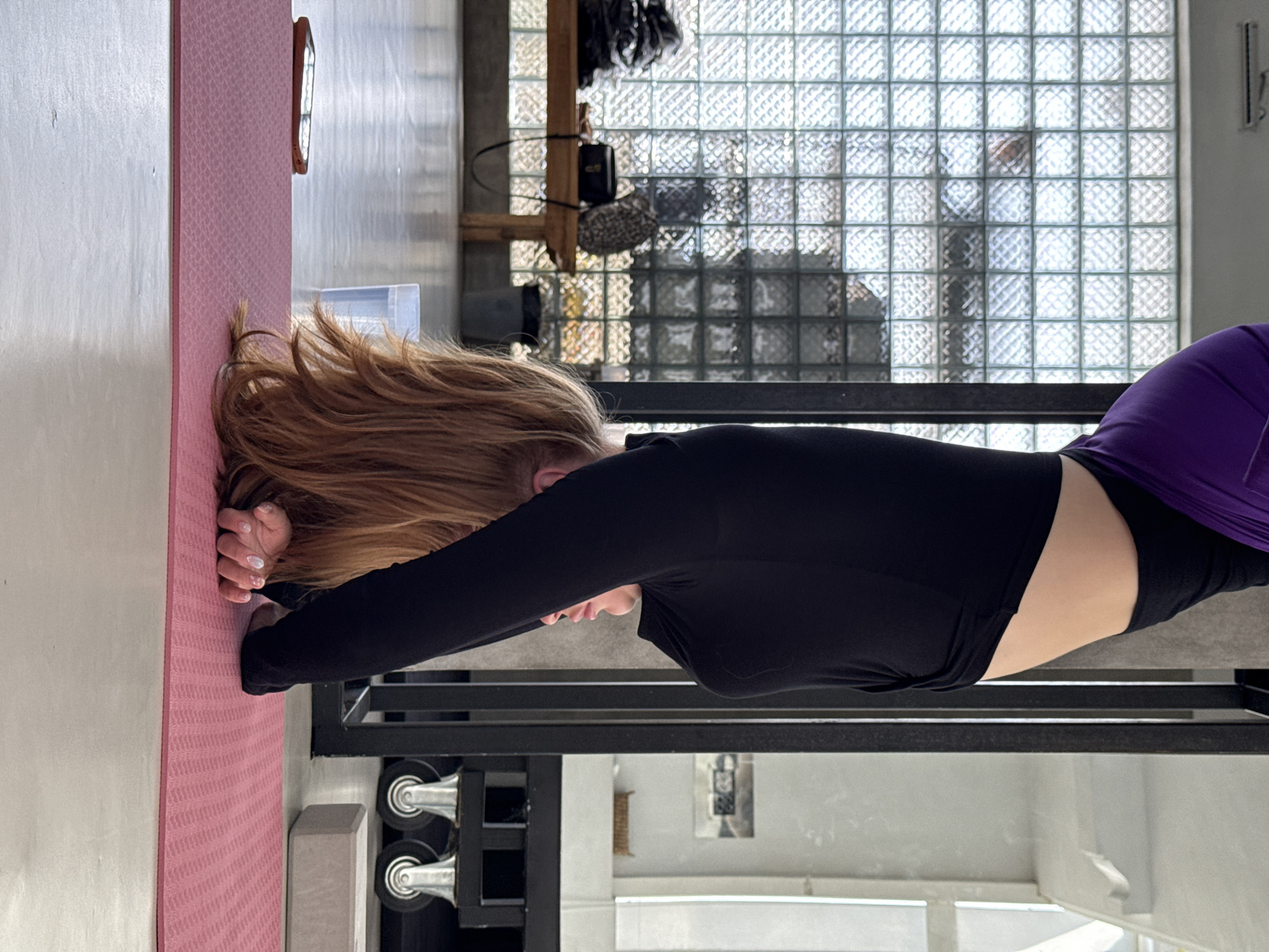 A woman is stretching on a pink yoga mat in a room. She is wearing a long-sleeved black top and purple leggings. Her hair is blonde and long, and she is positioned face-down on the mat with her arms extended in front of her. A yoga block sits next to the mat. Exercise equipment with wheels is visible on the floor near the yoga mat. The room has a window made of glass blocks. A metal structure is visible in the foreground. The location is Sukawati, Indonesia.