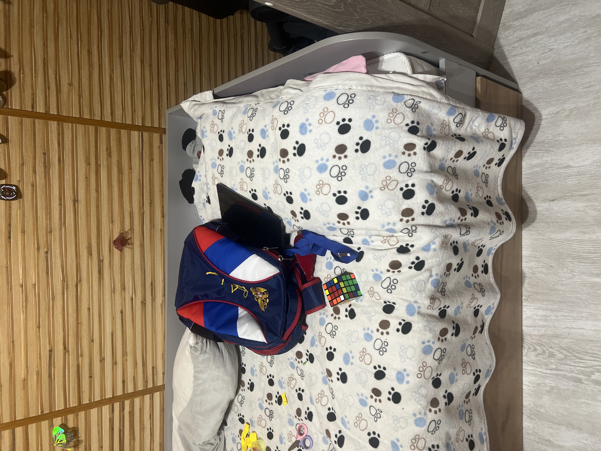 The image depicts a child's bed in a room with a bamboo-slatted wall. A backpack, a Rubik's cube, and a pair of scissors are scattered on a blanket adorned with paw prints. A pillow is also visible on the bed. The scene appears to be indoors, likely a bedroom. The location is identified as Surgut, Russia, although no specific geographic landmarks are visible in the image to confirm this. The lighting suggests it is daytime, possibly indoors with artificial lighting. There are no people visible in the image.