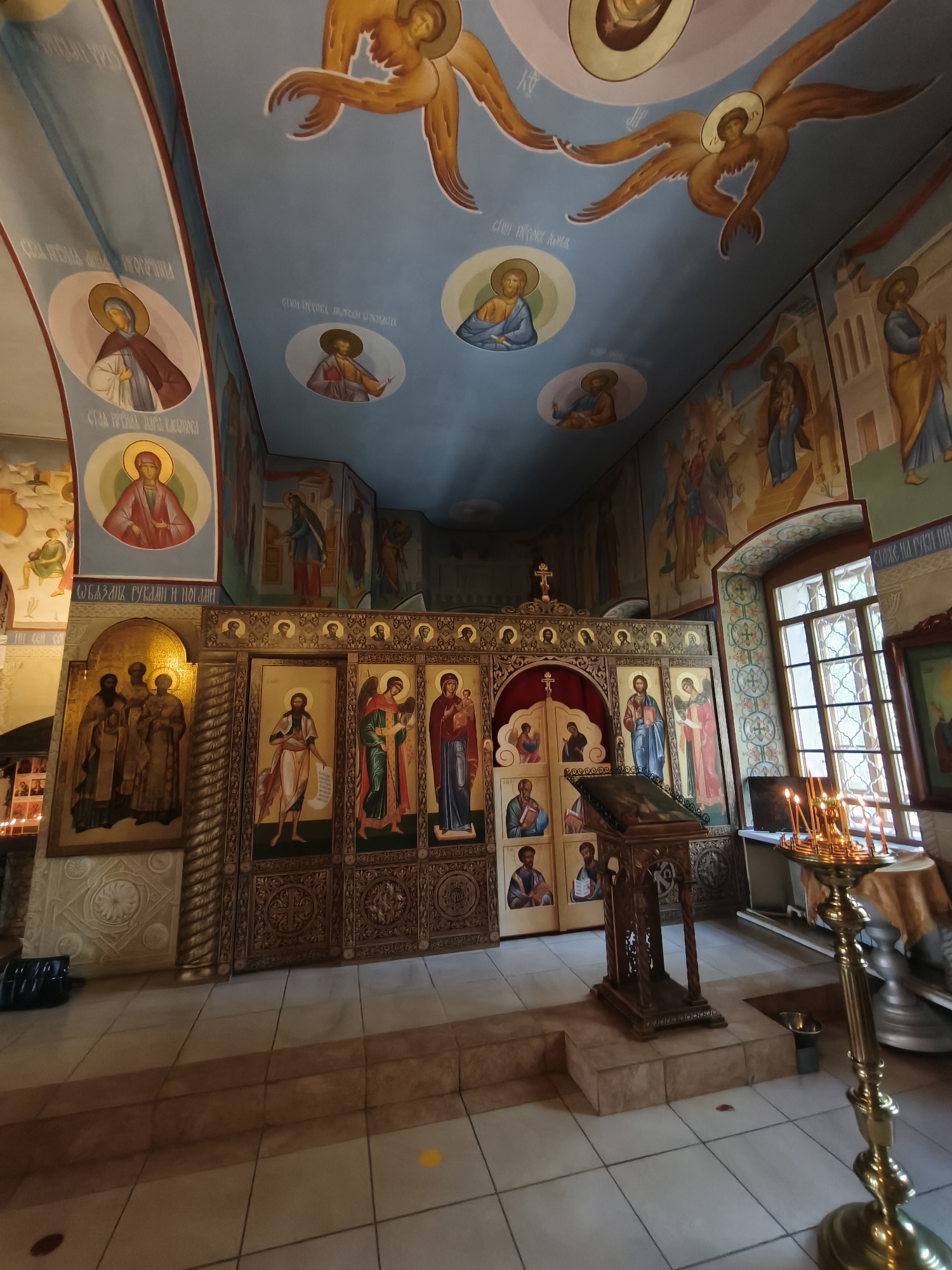 The image depicts the interior of an Orthodox church, specifically identified as being in the city of Kady, Russia. The scene is richly decorated with religious iconography, with numerous frescoes adorning the walls and ceiling. The ceiling features a blue expanse with painted depictions of angels with golden wings, and circular portraits of saints. The walls are covered in similar religious art, showing various figures in traditional Orthodox attire.
In the foreground, a prominent iconostasis, a wall of icons, separates the nave from the sanctuary. This structure is elaborately carved and painted, with multiple icons of saints and biblical scenes. To the right of the iconostasis, a wooden lectern stands, likely used for readings during services. In front of the lectern, a brass candelabra with lit candles illuminates the area.
The floor is tiled in a pattern of light and dark squares. To the left of the iconostasis, a large icon featuring a group of three figures in golden robes is visible. Further to the left, a wall with smaller framed pictures and a glimpse of what appears to be a candle holder suggests another area of the church. A window with intricate latticework is visible on the right side of the image, allowing natural light to enter the space. The overall impression is one of solemnity and religious devotion, characteristic of Orthodox Christian places of worship.