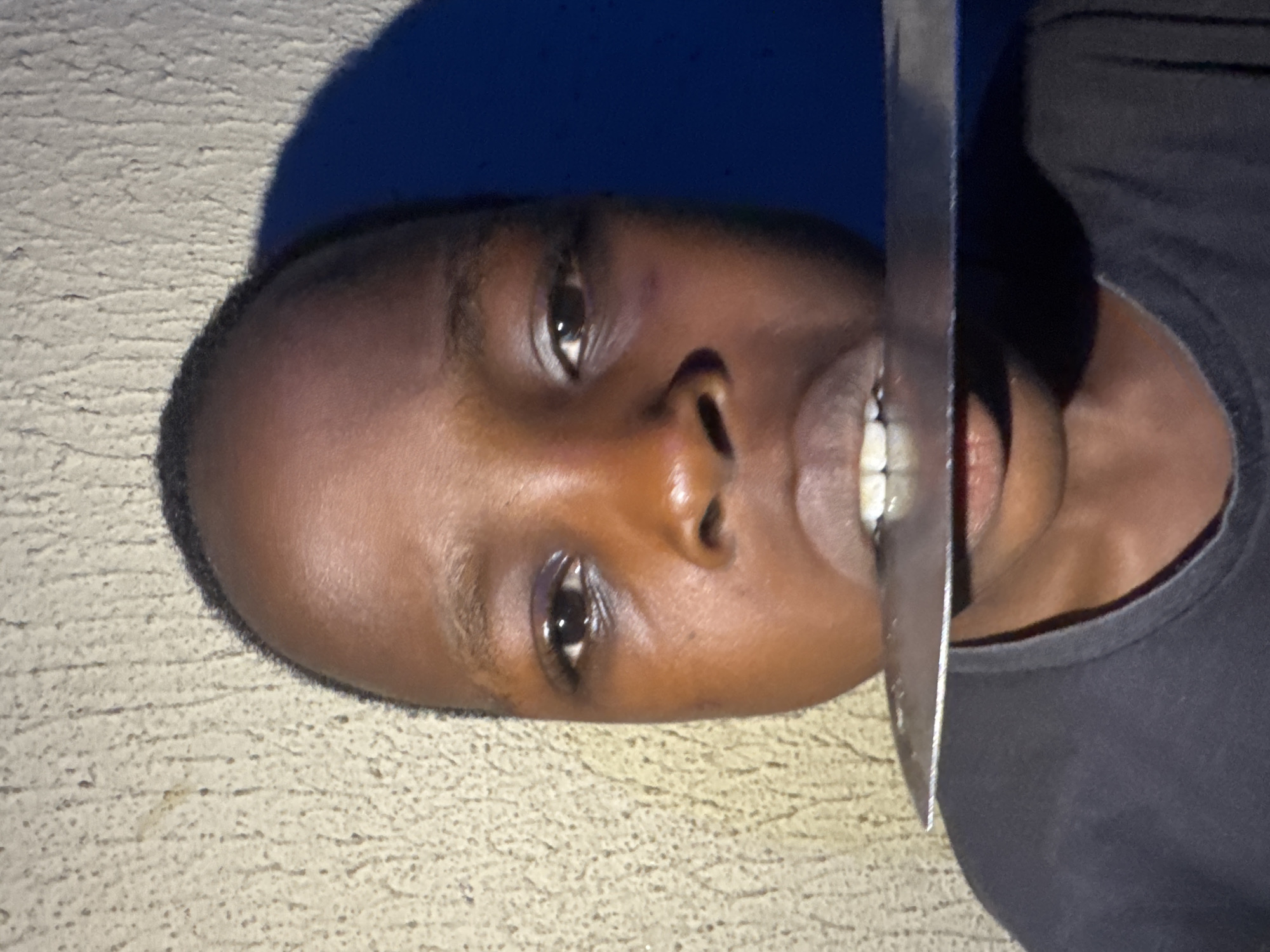 The image is a close-up portrait of a young person in Onitsha, Nigeria. The person's face is centered, and they are holding a knife horizontally across their mouth, with the blade inside their lips. The background is a textured wall, with a shadow cast on the right side of the frame. The lighting is harsh, creating strong contrasts. The young person's expression is neutral.