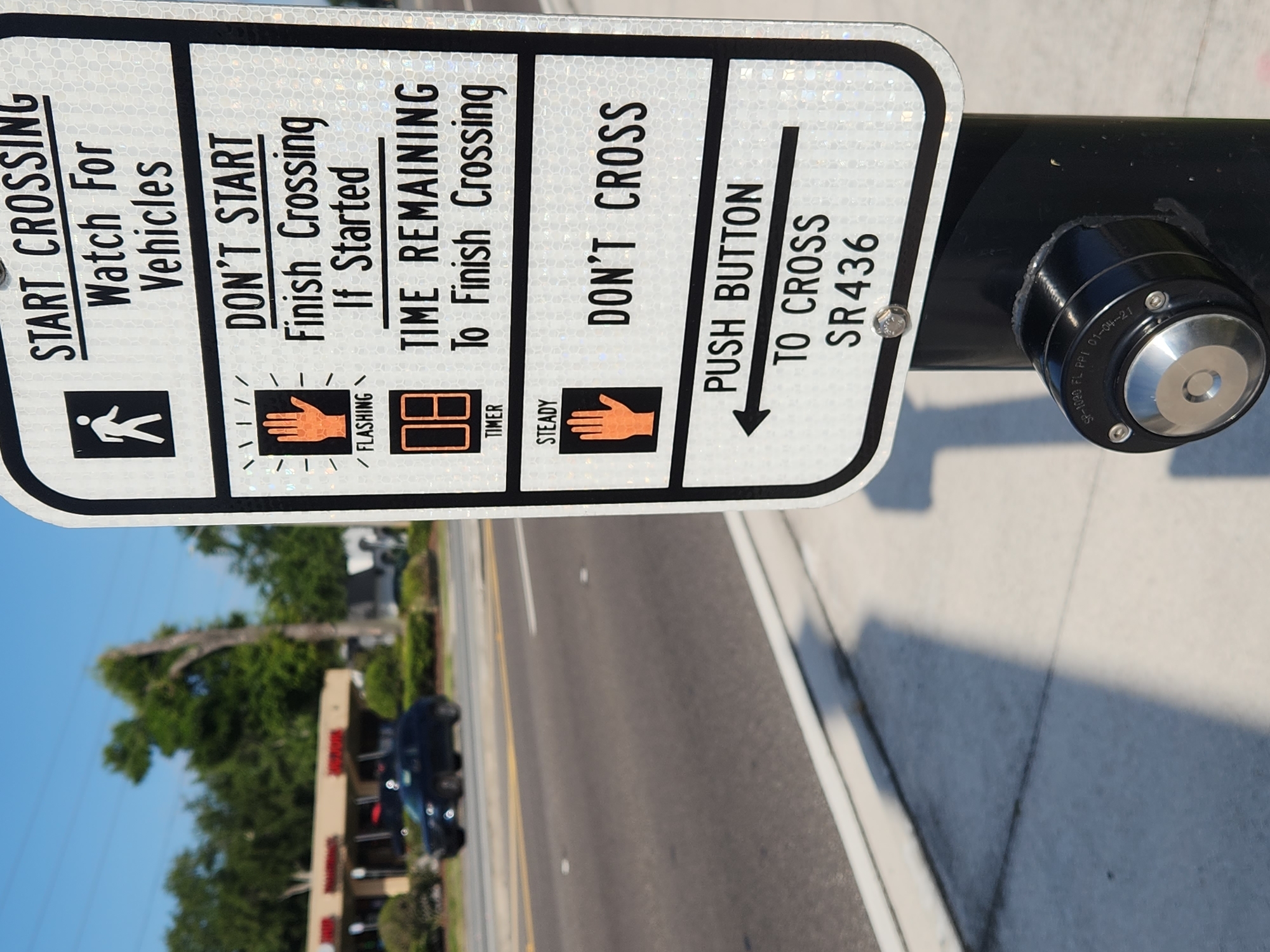 The image shows a pedestrian crossing signal sign and its associated push button, located on SR 436 in Altamonte Springs, United States of America. The sign is white with black lettering and reflective elements. It displays instructions for pedestrians, indicating when to start crossing, what to do if the light is flashing or steady, and when to press the push button. The push button is circular, made of metal, and located at the base of the pole supporting the sign. In the background, a dark-colored SUV is visible driving on the road, and businesses and trees line the side of the street. The sky is clear and blue, suggesting a sunny day. The overall scene depicts a typical urban environment with infrastructure designed for pedestrian safety.