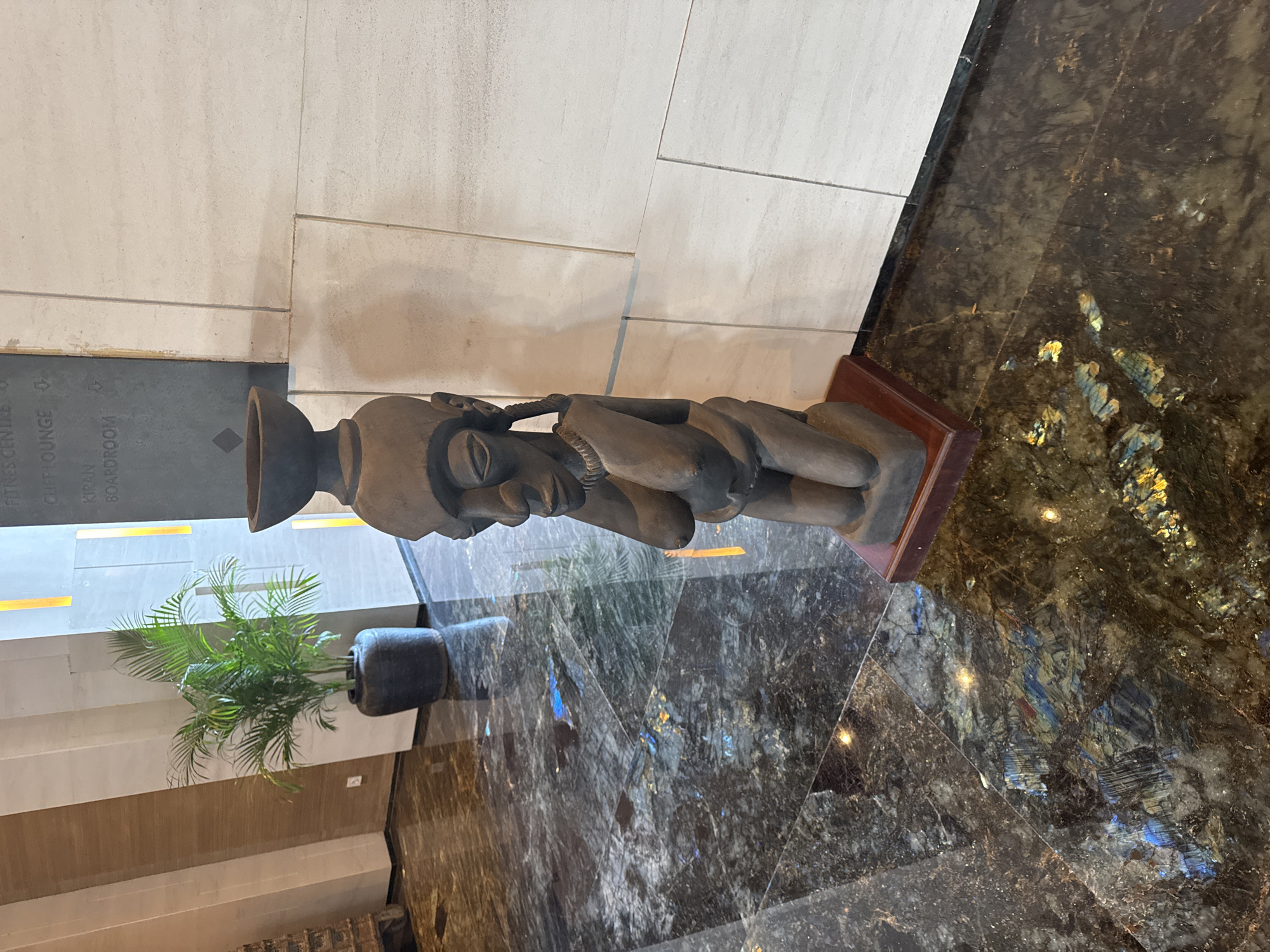 The image depicts an indoor scene in Kuta Selatan, Indonesia, featuring a carved wooden sculpture displayed on a wooden base. The sculpture is located near a potted plant and a stone wall, alongside a sign that lists "FITNESS CENTRE," "CLIFF LOUNGE," and "KIRAN BOARDROOM." The sculpture and plant are positioned on a polished stone floor with a mosaic-like pattern. The lighting is artificial. No people are visible in the image.