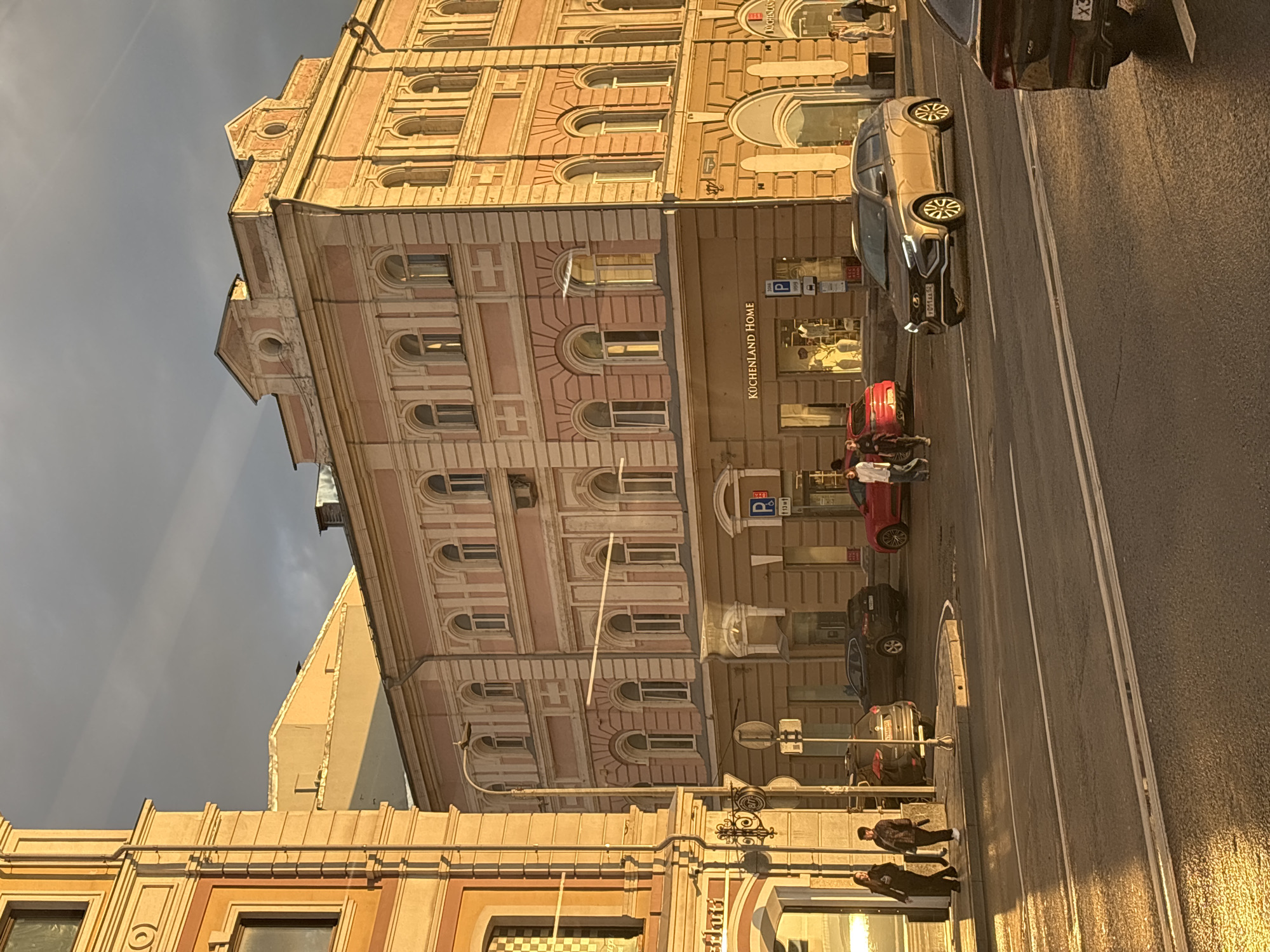 The image captures a street scene in Nizhny Novgorod, Russia, at what appears to be late afternoon or early evening, as indicated by the warm, low-angled sunlight. The focus is on a row of multi-story buildings lining a street. The buildings exhibit classical architectural styles with decorative facades, featuring arched windows, cornices, and mansard roofs. One building has a light pink facade with cream-colored detailing, and another to its left is a vibrant orange-yellow.
Several vehicles are present on the street, including a gold-colored car, a black SUV, and a red car. Two individuals are walking on the sidewalk in the foreground, and a few more people are visible further down the street near shop entrances. One shop clearly displays the name "KÜCHENLAND HOME". The sky above is partly cloudy with some streaks of light. The street and sidewalks appear wet, reflecting the sunlight. A street lamp is visible in the mid-ground. The overall impression is of a bustling urban environment bathed in the golden light of the setting sun.