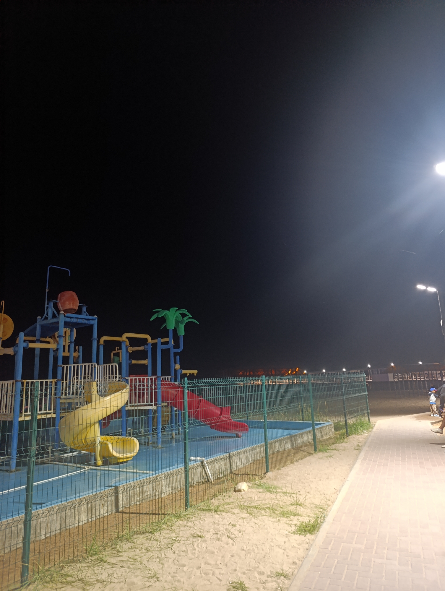 The image shows a playground at night in Bosteri, Kyrgyzstan. A colorful playground structure with blue, yellow, and red elements is visible, featuring slides and water features. The playground is enclosed by a green metal fence. To the right, a paved walkway runs alongside the sandy ground, and a lamppost illuminates the area. Several people are visible on the walkway in the distance, some appearing to be children and adults. The sky is dark, with a bright light source, likely a street lamp, casting a strong glare in the upper right corner of the image.