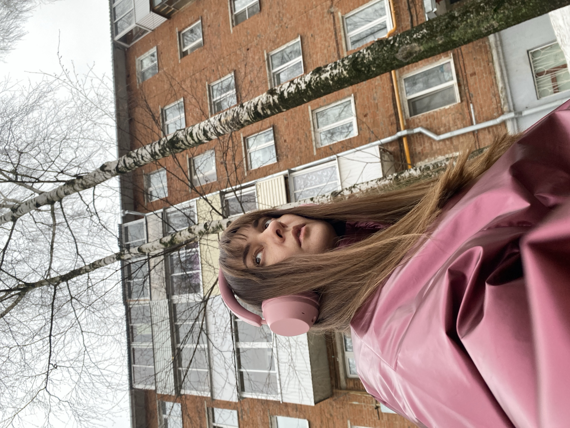 A low-angle, off-center shot captures a woman with long brown hair and bangs wearing pink headphones and a shiny pink jacket. She is looking up at the camera with a neutral expression. A tree branch with white bark and bare twigs diagonally crosses the upper portion of the frame. Behind the woman and tree, a multi-story brick building with rows of windows is visible. The location appears to be Votkinsk, Russia, based on architectural and environmental characteristics commonly found in urban areas of that region. The sky is overcast, suggesting a cold or winter day.