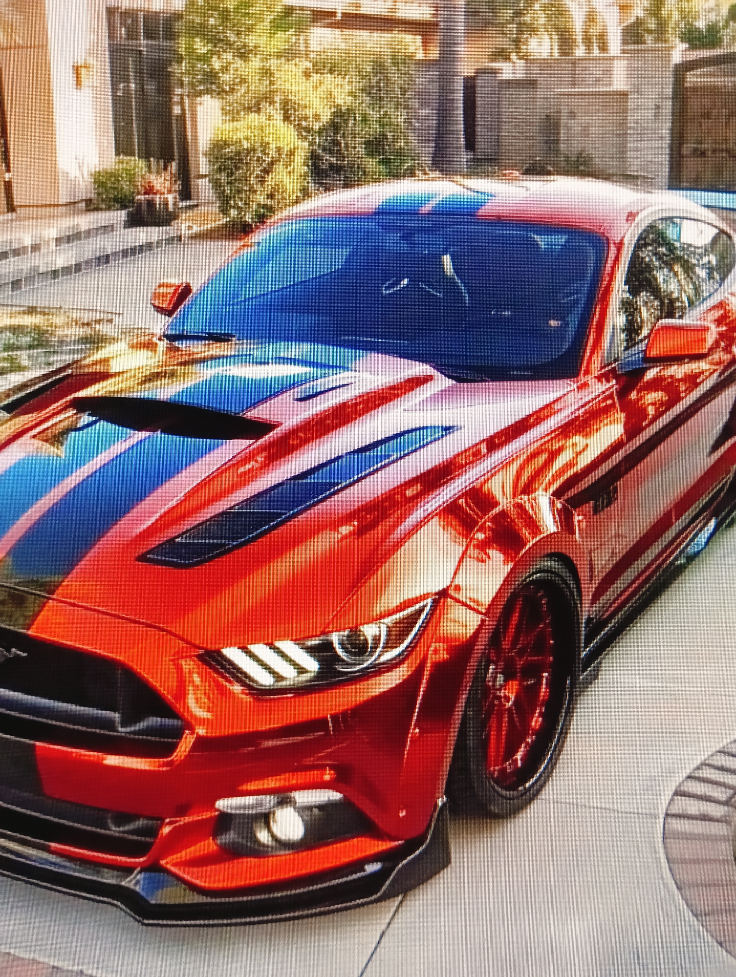 The image shows a custom orange Ford Mustang with black racing stripes parked in a driveway. The car has a wide body kit, aftermarket wheels, and a lowered suspension. The background features a modern house with a manicured lawn and palm trees. The driveway appears to be made of stone pavers. The lighting suggests it is daytime, possibly late afternoon, given the warm tones. There are no people visible in the image. The scene depicts a luxury vehicle in a residential setting. There is no information within the image to suggest a connection to the city of Sluck, Belarus.