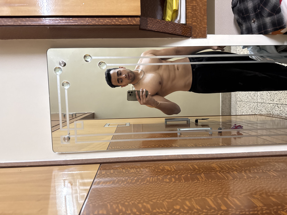 A muscular man with short dark hair, wearing only black sweatpants, a silver chain, and a thin gold bracelet, is seen in a mirror selfie taken indoors in Kolkata, India. He holds a grey smartphone in his right hand, capturing his reflection in a rectangular wall-mounted mirror. The mirror features etched geometric patterns and four circular decorative elements. The man exhibits visibly defined abdominal muscles and a neutral facial expression with slightly raised eyebrows and a partially open mouth. To the left of the mirror is a light brown wooden wardrobe with a textured, patterned surface. The mirror's reflection reveals a light-colored wall, a second wooden cabinet with two silver handles, and a light-colored speckled tiled floor. To the right of the mirror, a darker brown wooden shelving unit contains a yellow object and a stack of clothing, including a plaid shirt. The scene is evenly lit by indoor lighting.