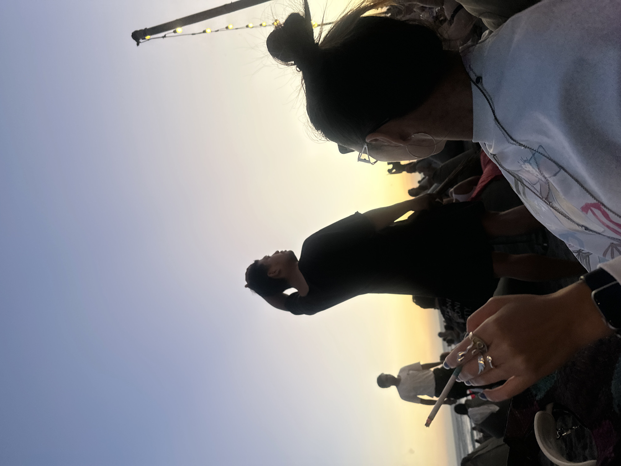 The image captures an outdoor scene at sunset or sunrise, with a prominent golden glow on the horizon transitioning into a soft blue sky. The photo is taken from a low angle, looking up towards the sky and the people present.
In the immediate foreground, to the right, a person is visible from the chest up, looking downwards. Their dark hair is tied in a bun, and they wear glasses with distinct geometric frames. They are dressed in a light-colored top, possibly light blue or grey, with a faint, unreadable design. Their right hand, adorned with multiple rings (one appearing distinctive, possibly frog-shaped) and blue nail polish, holds a lit cigarette or joint. A dark watch is visible on their left wrist.
Further back, in the mid-ground, several individuals are silhouetted against the bright sky. One person, wearing a dark top, stands with their back mostly to the viewer, their head tilted slightly upwards and an arm raised towards their head or neck area, appearing to be observing the sky. Behind them, another person in a lighter-colored shirt is also visible as a silhouette. Other indistinct figures are present in the deeper background.
In the upper left corner, a dark pole or beam extends into the frame, from which a string of small, glowing yellow lights is suspended. The horizon line suggests a vast open space, possibly a beach or coastal area, with what appears to be the ocean beneath the sky's glow. The overall atmosphere is serene and contemplative, characteristic of an evening or morning gathering in North Kuta, Indonesia. No discernible text is visible in the image.