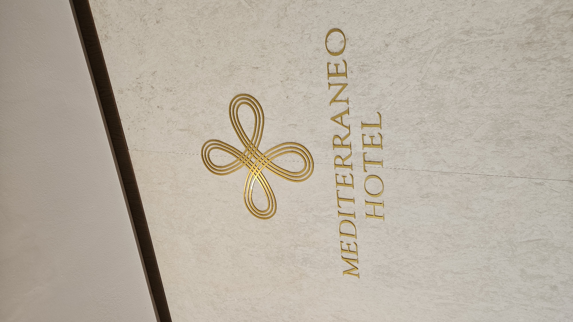 The image shows a close-up of the sign for the Mediterraneo Hotel. The sign is set on a beige, textured surface. To the left of the text is a gold-colored stylized floral symbol. The hotel name, "MEDITERRANEO HOTEL," is printed vertically in gold, sans-serif block letters. The edge of a ceiling is visible on the left with a dark wood trim. The hotel is located in the city of Chersonisos Municipal Unit, Greece.