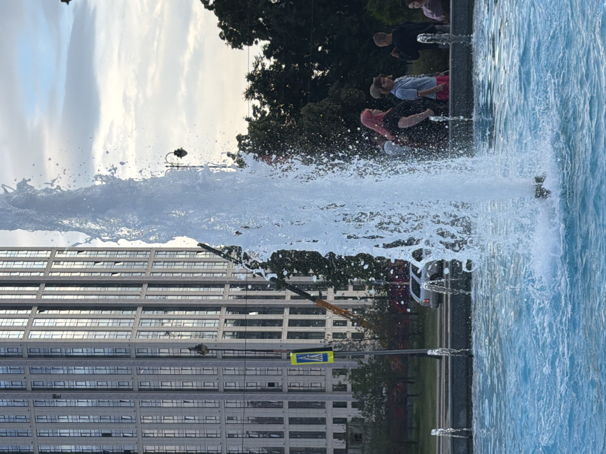 In the city of Unknown, Unknown, a fountain sprays water in the foreground. In the background, a tall, modern building stands with many windows. A street with cars and trees is visible between the fountain and the building. Several people are gathered near the fountain, some with brightly colored hair. The sky is cloudy.