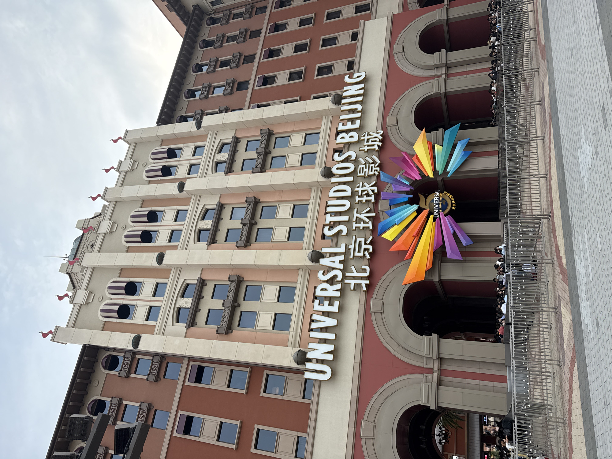 The image shows the entrance of Universal Studios Beijing, located in the Tongzhou District of China. The building has a multi-story facade with red and tan coloring, arched openings, and many windows. The Universal Studios Beijing logo is prominently displayed, featuring the words "UNIVERSAL STUDIOS BEIJING" in English and Chinese characters, along with a colorful, sun-like symbol. A crowd of people is visible near the entrance. A metal fence separates them from the entrance area. The sky is visible in the upper left corner.