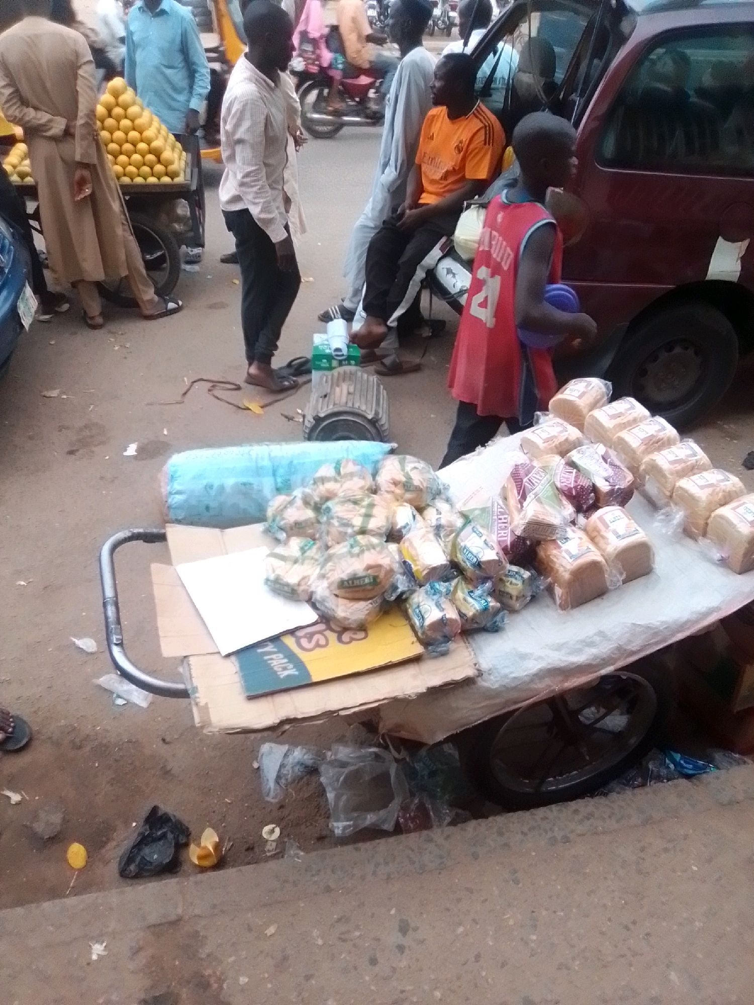 The image depicts a street scene in Arawa, Nigeria, likely a market area given the presence of vendors and goods for sale. In the foreground, a cart is laden with numerous loaves of bread, most of which are individually packaged in plastic. A large blue plastic-wrapped object, possibly insulation or a similar material, is also on the cart. The cart itself appears to be made of metal and has a wheel visible underneath. To the left of the cart, a pile of oranges is displayed on another cart, suggesting a fruit vendor.
Several people are present. A man in a light-colored collared shirt and dark pants stands near the oranges, facing away from the camera. Another man, wearing an orange t-shirt, is seated. A young boy in a red sleeveless jersey with the number "24" and "DAVID" on it is also visible, holding a blue object. Various other individuals are partially visible in the background, some near a maroon vehicle. The ground is paved with concrete, and there is scattered debris, including plastic bags and what appear to be discarded food items. The overall impression is one of a busy, outdoor marketplace.