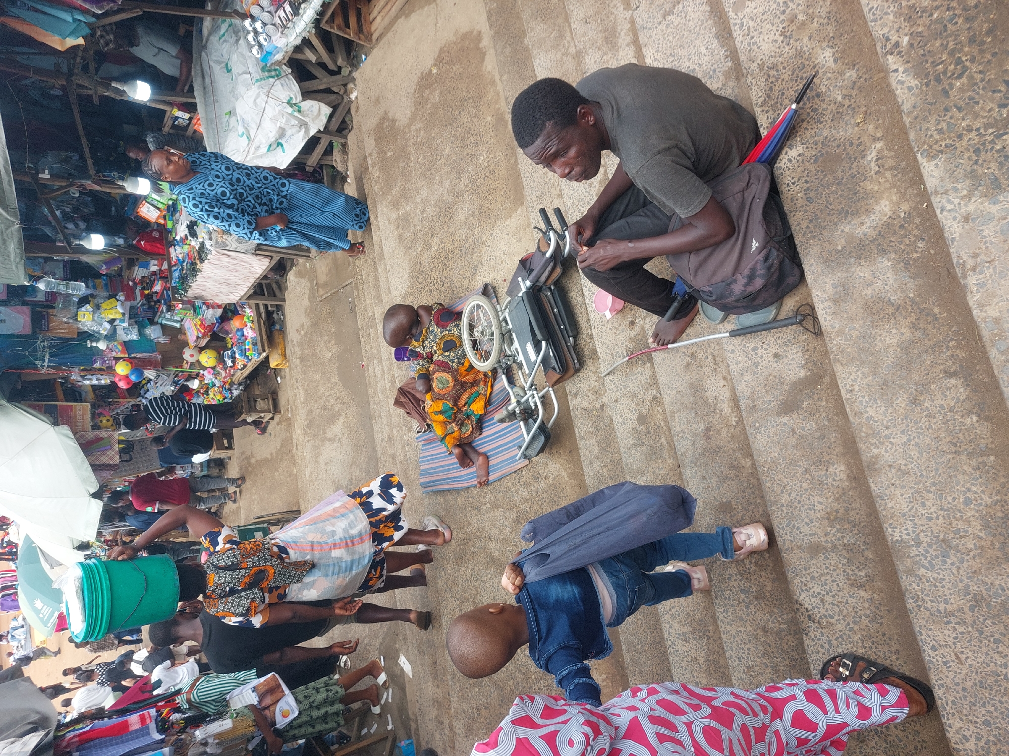 The image shows a street scene in Dar es-Salaam, Tanzania.
A man sits on concrete steps next to a folded wheelchair and a child seated on a blanket. The man is dressed in a grey shirt and dark pants and is barefoot, and a backpack sits behind him. A red, white, and blue umbrella is leaned against the steps nearby. The child seated on the blanket is wearing colorful African fabric clothing.
Pedestrians are walking nearby, including a woman in a white and blue dress, a person carrying a green bucket on their head, a person wearing striped clothes, and others.
The scene appears to be a market or commercial area, with stalls, goods, and lighting fixtures visible in the background.