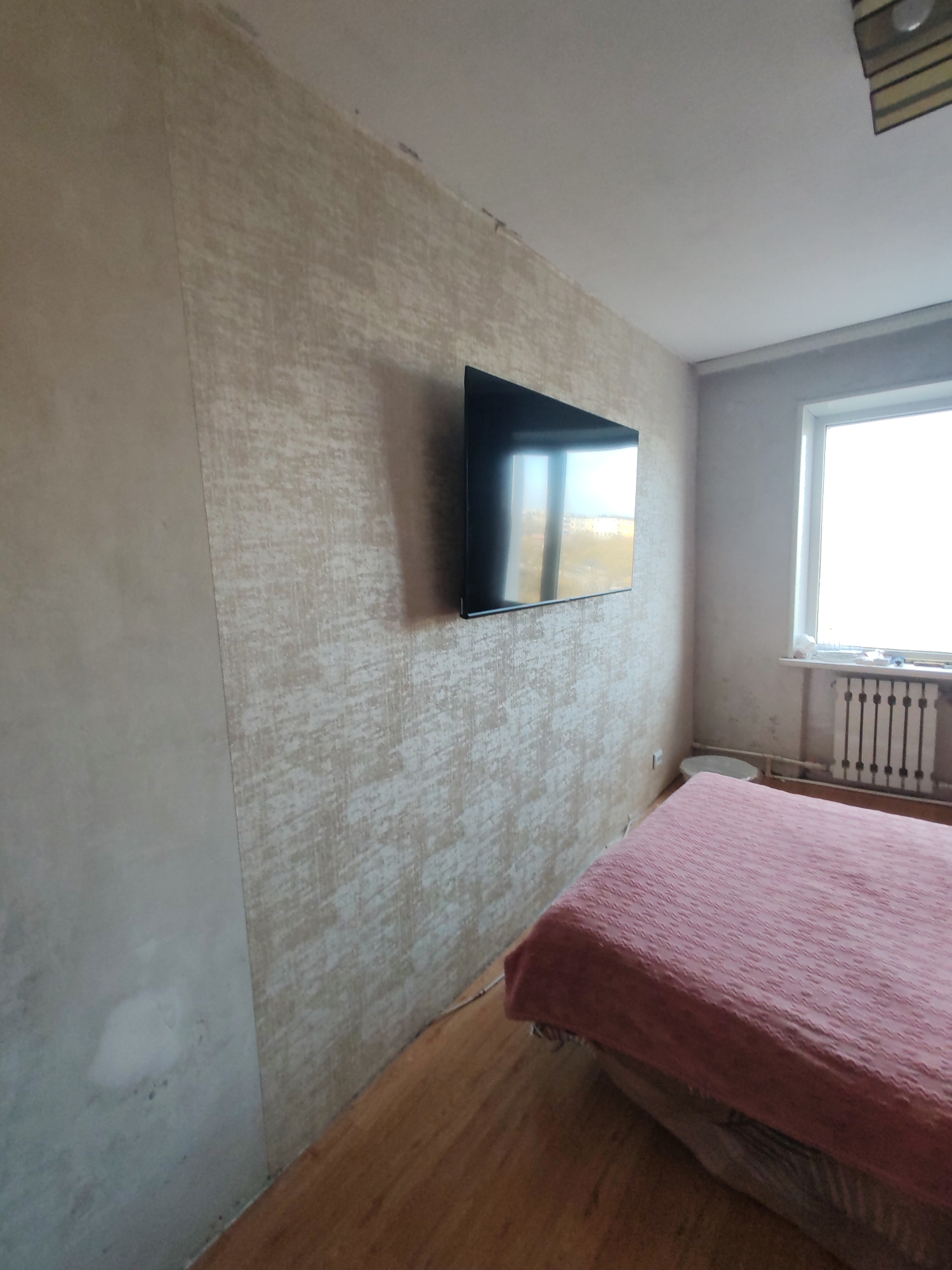 The image shows a bedroom in a building located in Petropavlovsk-Kamchatsky, Russia. The room is modestly furnished and appears to be undergoing renovation or in need of repair. The walls are covered with two different types of wallpaper. The wall on the left appears to have remnants of old wallpaper or unfinished painting. The wall on the right, behind the bed, has a patterned, beige wallpaper. A flat-screen television is mounted on the patterned wall.
The room contains a bed with a red-pink bedspread. The flooring is made of wooden planks.
In the distance is a window with a radiator beneath it. The lighting in the room suggests it is daytime. No people are visible.