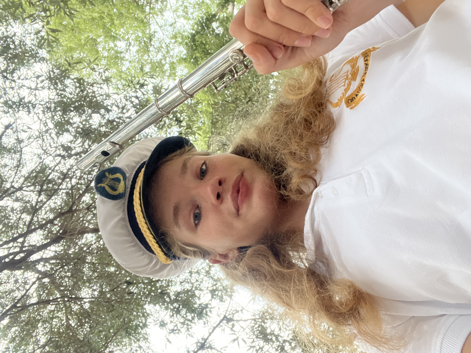 This is a selfie of a young person holding a flute. The person is wearing a white polo shirt with an embroidered emblem on the left side of the chest and a white cap with a dark band and gold trim. The cap has a similar emblem to the shirt. The person has blonde, curly hair and is looking at the camera with light blue eyes. The background consists of out-of-focus green leaves and branches, suggesting an outdoor setting among trees. The image is oriented sideways. The time of day appears to be daytime, and the weather seems fair, given the natural light. There is no visible text other than the embroidered emblems on the clothing, which are not legible.