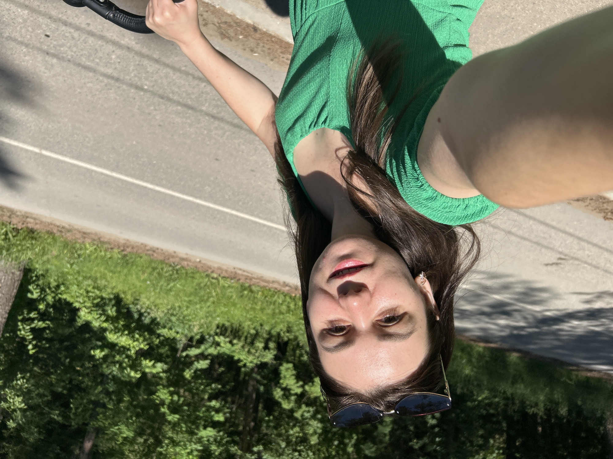 A young woman with dark hair and sunglasses pushed up on her head is taking a selfie from her perspective. She is wearing a green textured dress and has a mole on her right arm. She is outdoors on a paved road, with lush green trees and foliage lining the side of the road. The sun is shining, casting shadows on the road. The location appears to be a park or a rural road, consistent with the surroundings of Chaikovsky, Russia. The woman is holding what appears to be the handlebars of a bicycle or stroller.