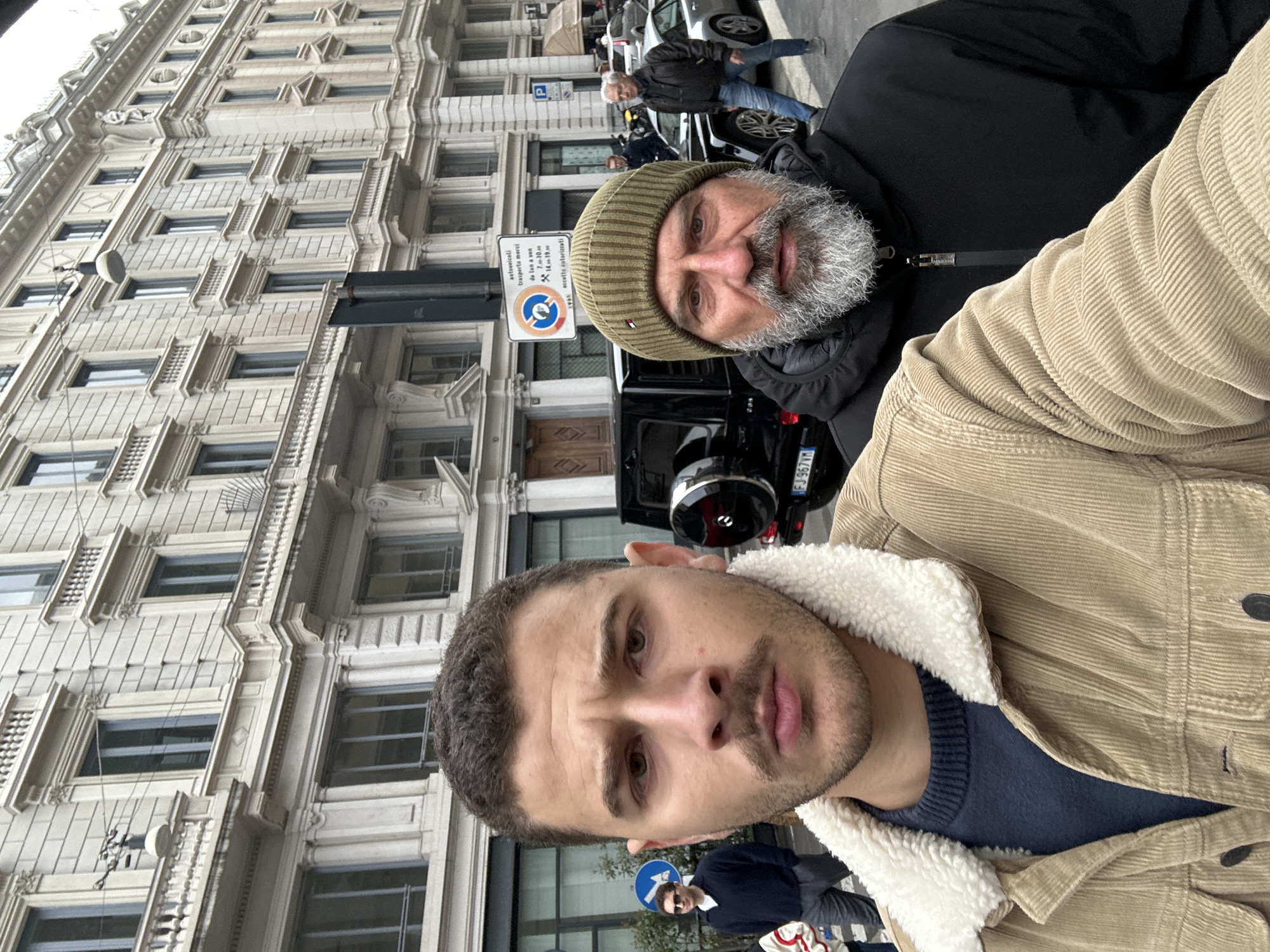The image captures a daytime street scene in central Milan, Italy, featuring two men in the foreground appearing to take a selfie, with an ornate building and other urban elements in the background.
The primary subject, positioned on the left and center of the foreground, is a young adult male with short, dark, curly hair and a mustache. He wears a navy blue crew-neck sweater under a light brown corduroy jacket with a white sherpa collar, and is looking directly at the camera with a neutral, slightly furrowed expression.
Immediately behind and to the right of the primary subject stands an older adult male with a full grey beard. He wears an olive green knit beanie and a black hooded puffer jacket, zipped, and is also looking directly at the camera with a neutral expression.
In the middle ground, a black Mercedes-Benz G-Class SUV (license plate FJ 967VM) is parked. Behind it, a silver Porsche is partially visible. A black pole displays a white rectangular street sign with Italian text: "autoveicoli trasporto merci da lun a ven 7-10 14-19 eccetto autorizzati" (commercial vehicles for goods transport from Mon to Fri 7-10, 14-19, except authorized), mounted below a circular "no stopping/standing" sign. To the left of the pole, a circular blue pedestrian sign with a white arrow points right. Other pedestrians, including a man in sunglasses on the far left and a man in a black jacket and jeans on the far right, are visible in the blurred background.
The background is dominated by a multi-story, classical-style building constructed from light-colored stone, featuring numerous rectangular windows, ornate decorative details, and balconies with balustrades. This architectural style and the specific Italian street signage are characteristic of central Milan, Italy, likely an affluent area such as the Quadrilatero della Moda or Corso Venezia. The lighting suggests an overcast day.