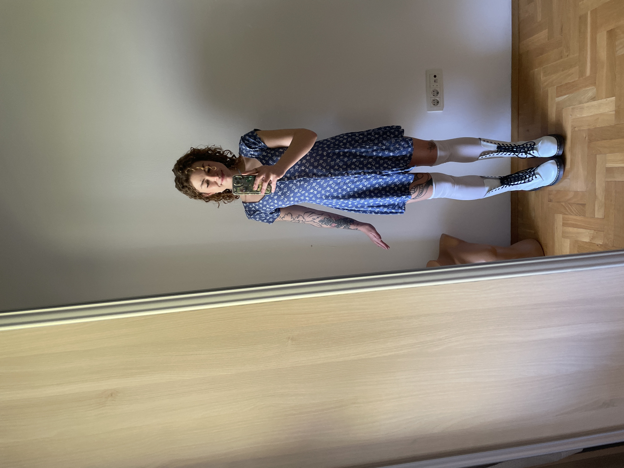 A young woman is reflected in a mirror. She is wearing a short, blue, floral dress, white knee-high socks, and white boots. She is holding a cell phone, apparently taking a picture of herself. The background includes a beige wall with an electrical outlet, a light wood-colored parquet floor, and a nude mannequin. The image does not contain information about Boreti, Montenegro.