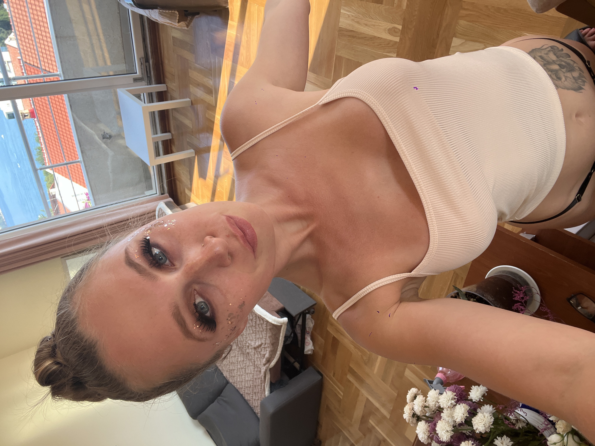 A woman with blonde hair styled in a bun is taking a selfie in what appears to be an apartment with a view of the sea and red-tiled roofs in Bečići, Montenegro. She is wearing a light-colored ribbed tank top and has glitter on her face and makeup. In the background, there is a couch, a small white table, and a balcony door opening to the outdoor view. A bouquet of flowers is visible on a table in the foreground, along with a potted plant. A tattoo of an animal is visible on her stomach.