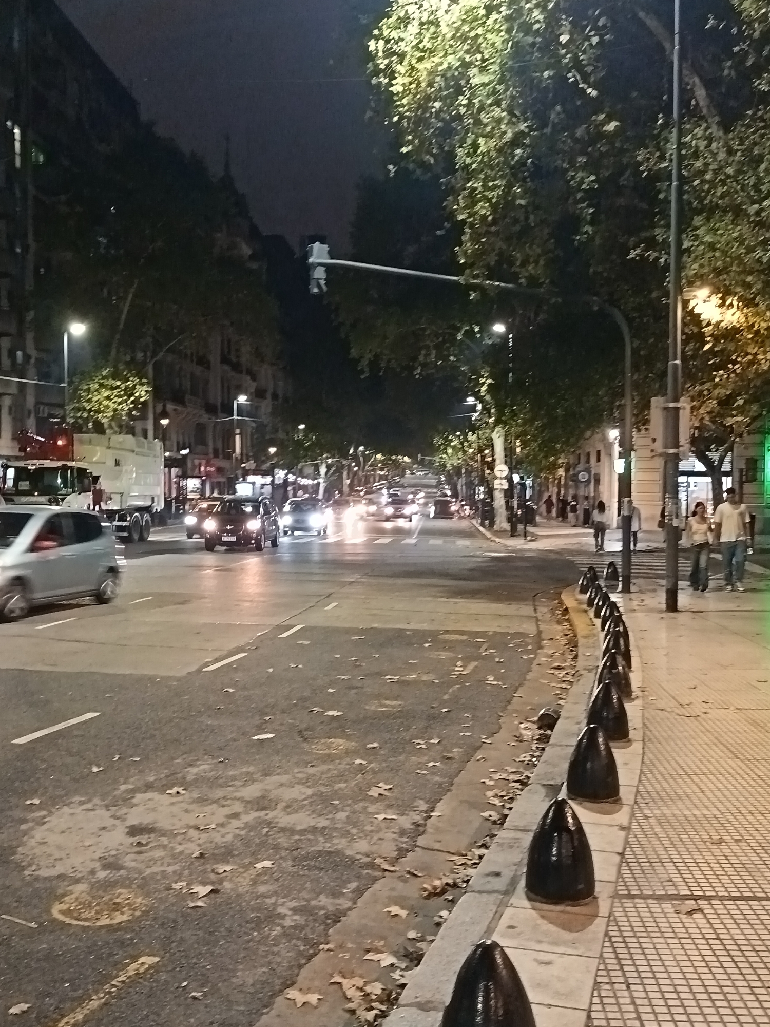 The image depicts a nighttime street scene in Buenos Aires, Argentina. A wide avenue with multiple lanes is visible, with car traffic heading towards the vanishing point in the distance. On the left, a large white truck is parked near the curb, and cars with headlights illuminated move along the avenue. The road surface is marked with lane lines and shows scattered fallen leaves.
On the right side of the image, a sidewalk is tiled in a checkered pattern. A row of dark, cone-shaped bollards lines the edge of the sidewalk, separating it from the street. Two people are walking together along the sidewalk, while another individual stands near a traffic light pole. Trees with illuminated foliage line both sides of the avenue, and buildings are visible in the background. Streetlights provide illumination.