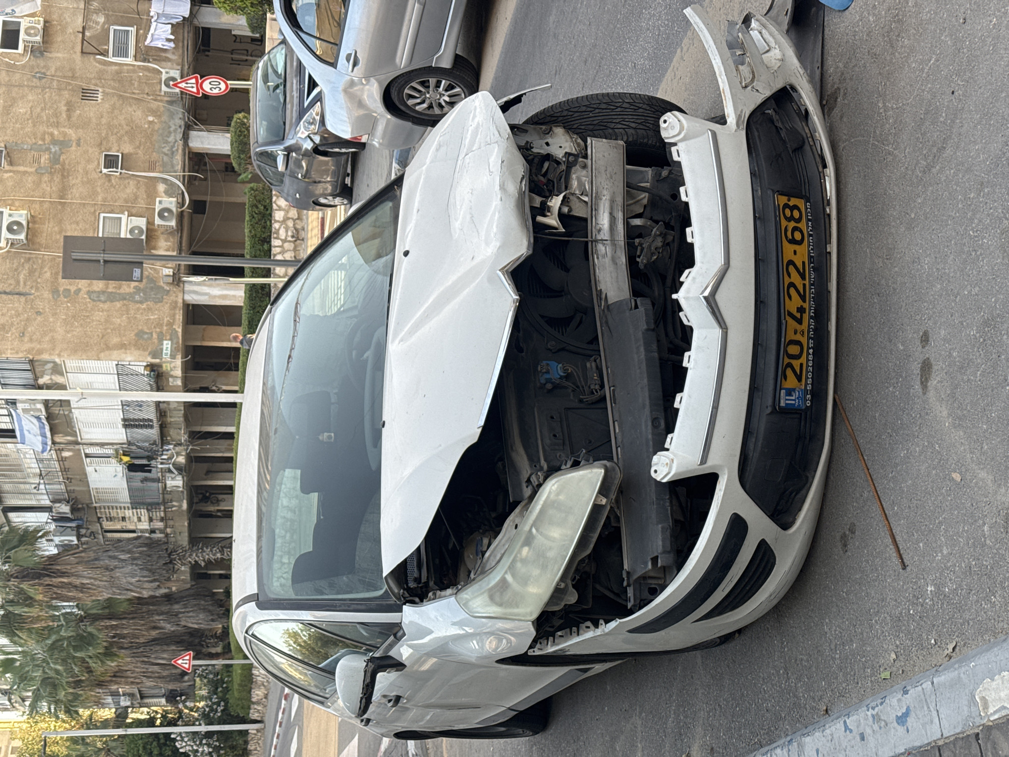 The image depicts a severely damaged white car, a Citroen, on a street in what appears to be an urban setting. The front end of the vehicle is significantly crushed, with the hood crumpled and the front bumper detached. The engine compartment is exposed, revealing internal components. The car's license plate is visible, bearing the number 20-422-68, which identifies it as registered in Israel. The image includes the words "מכון אלון" which is the name of a car testing company in Israel.
In the background, there are residential buildings with multiple air conditioning units attached to the facades. A street sign is also visible, indicating a speed limit of 30 kilometers per hour. Another car, silver in color, is parked nearby. The general environment suggests the location is in a residential or commercial area of a city. The context strongly indicates that the scene is in Bat Yam, Israel.