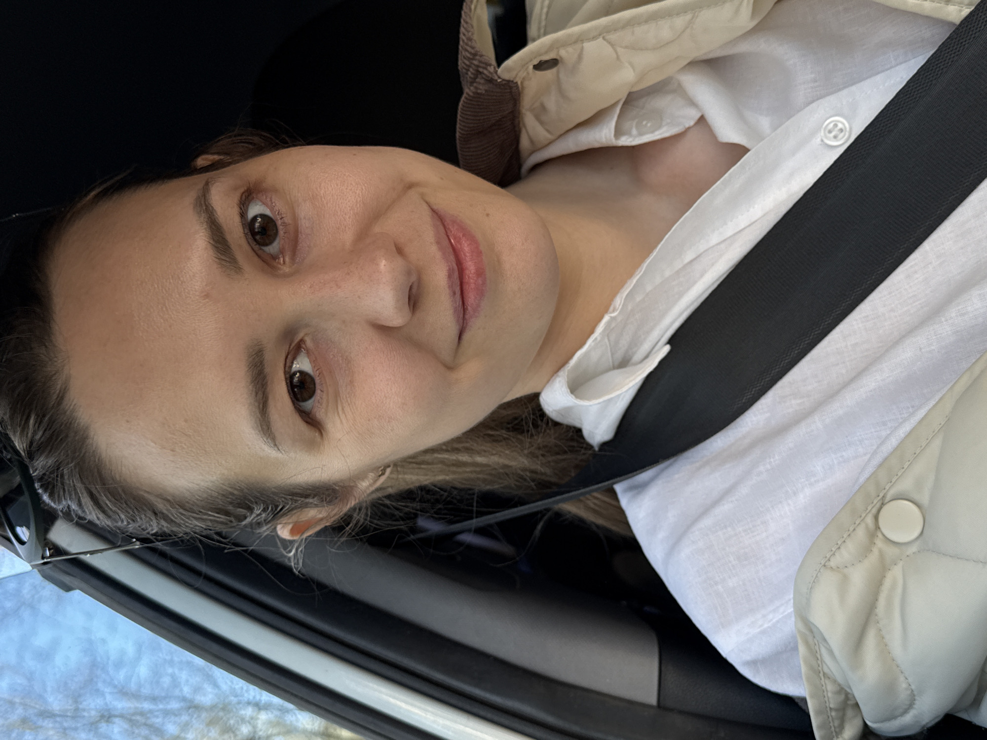 A woman is in a car wearing a white shirt, a quilted jacket, and a seatbelt. It is difficult to determine the exact location within Chaikovsky, Russia from the image provided.