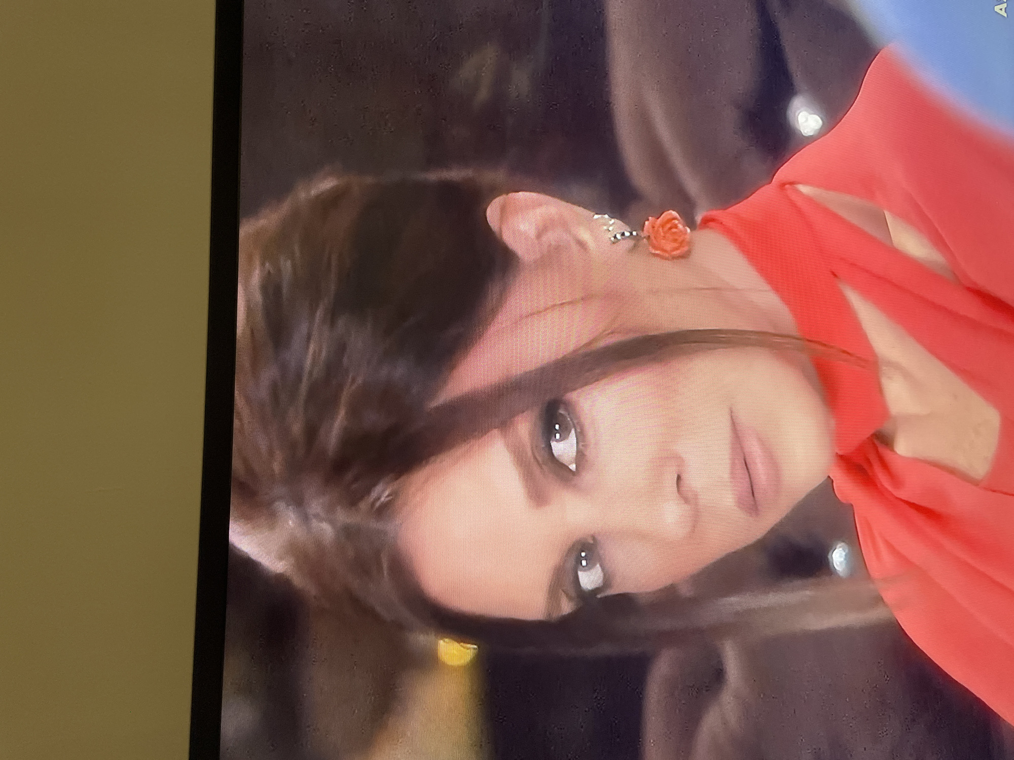 The image is a close-up, slightly blurry, headshot of a woman with dark brown hair styled up and to the side, with strands framing her face. She is wearing a bright coral-colored top with a high neckline and cut-out details. Her makeup is noticeable, with smoky eye shadow and light lipstick. She has a single coral rose earring on her visible left ear. The background is out of focus and appears to be dimly lit with some indistinct shapes that might suggest furniture or other people. There is no identifiable information in the image or its crops to suggest a location in Kadıköy, Turkey.