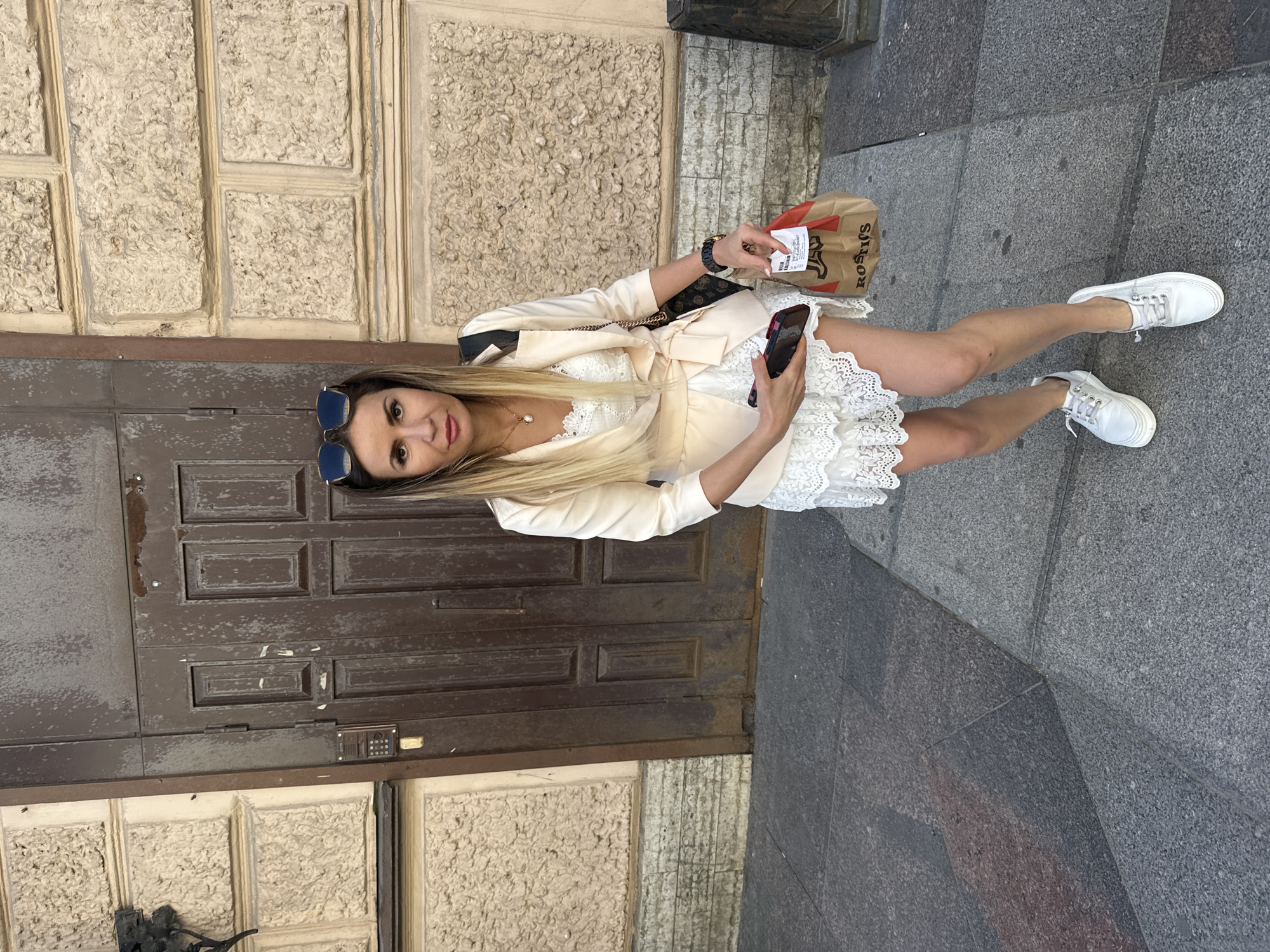 A woman stands on a paved walkway in front of a large, weathered brown wooden door set in a building with a textured, beige stone facade. She is wearing a white lace dress and a light-colored jacket tied at the waist. She has blonde hair and is wearing sunglasses on top of her head. In her left hand, she holds a dark smartphone, and in her right hand, she carries a paper bag with the "Rostik's" logo. She is also wearing white sneakers and a watch. The image appears to be taken during daylight, possibly in a historic district of Saint Petersburg, Russia, given the architectural style of the building.