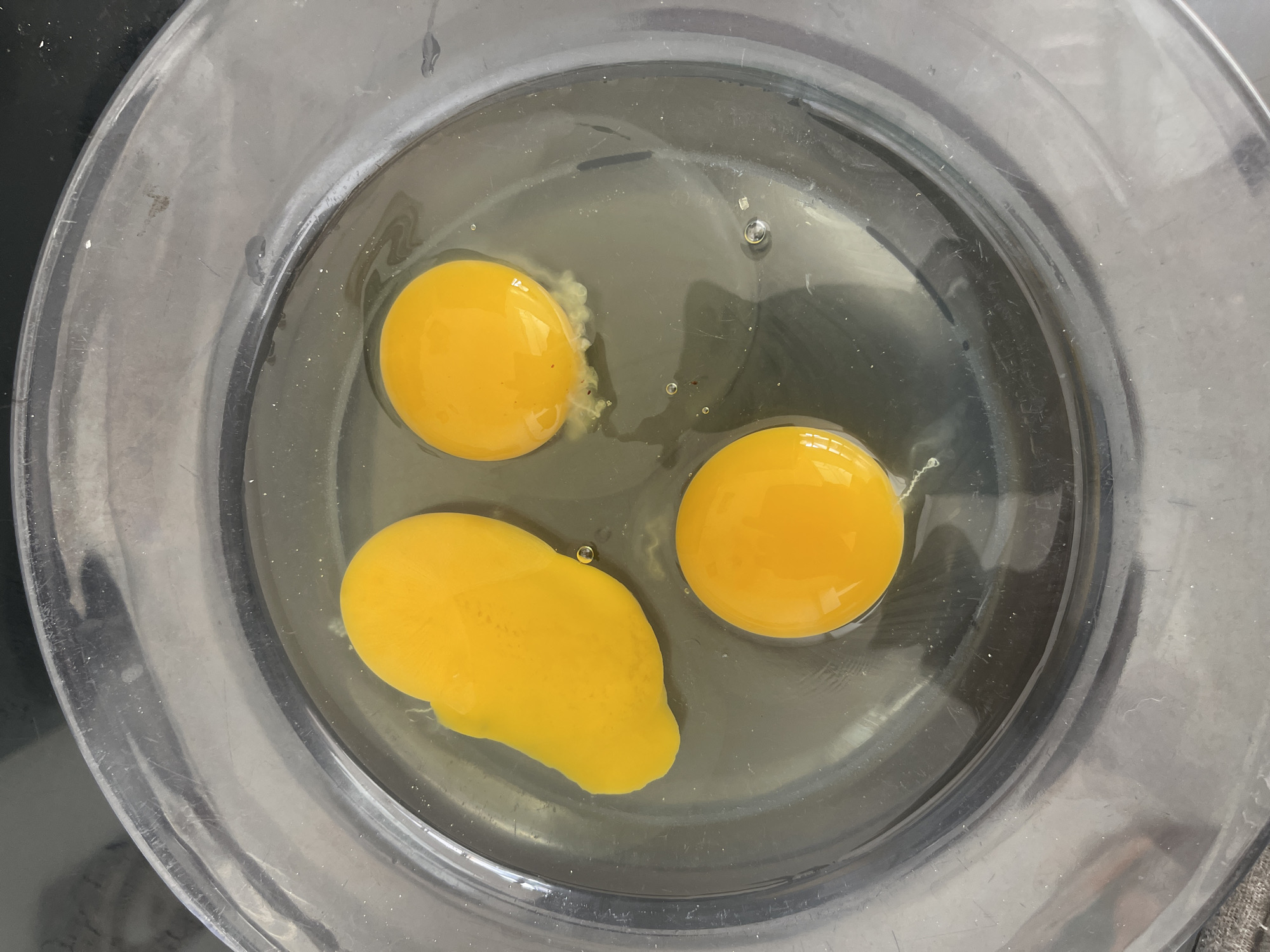 Three egg yolks and one egg white are in a clear glass bowl. The bowl is filled with a small amount of liquid, likely water. The bowl is on a dark, speckled surface. The eggs are bright yellow and appear to be fresh. One yolk is elongated, while the other two are round. There are a few small bubbles in the liquid. The image is taken from a top-down perspective.