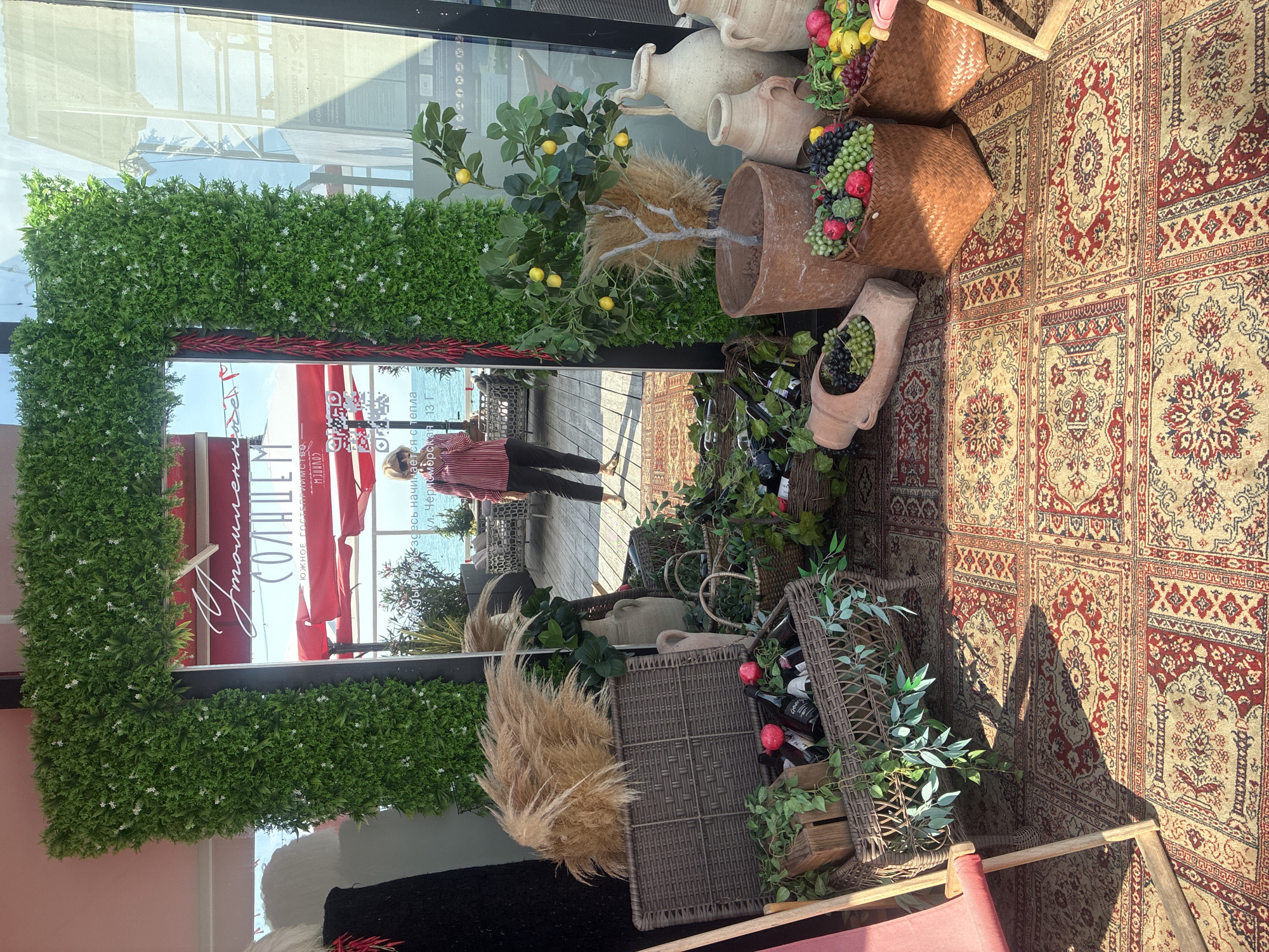 This image depicts an outdoor scene, likely a cafe or restaurant terrace in Sochi, Russia, given the visible text. The setting is decorated with artificial greenery, ceramic vases, woven baskets filled with faux fruits (grapes, lemons, pomegranates), and pampas grass. A large mirror is positioned vertically, reflecting a person in a striped shirt walking towards it and a glimpse of the sea and red awnings in the background. The ground is covered with a richly patterned, oriental-style rug. The sun appears to be shining, casting shadows on the rug, suggesting it's daytime, possibly afternoon. The overall impression is one of a charming, Mediterranean-inspired outdoor seating area. Visible text includes "СОЛНЦЕ" (Sun), "ЮЖНОЕ ГОСТЕПРИИМСТВО" (Southern Hospitality), "Кждый день здесь начинается с тепла" (Every day starts with warmth here), and an address "ул. Черноморская 13 Г" (Chernomorskaya St. 13 G).