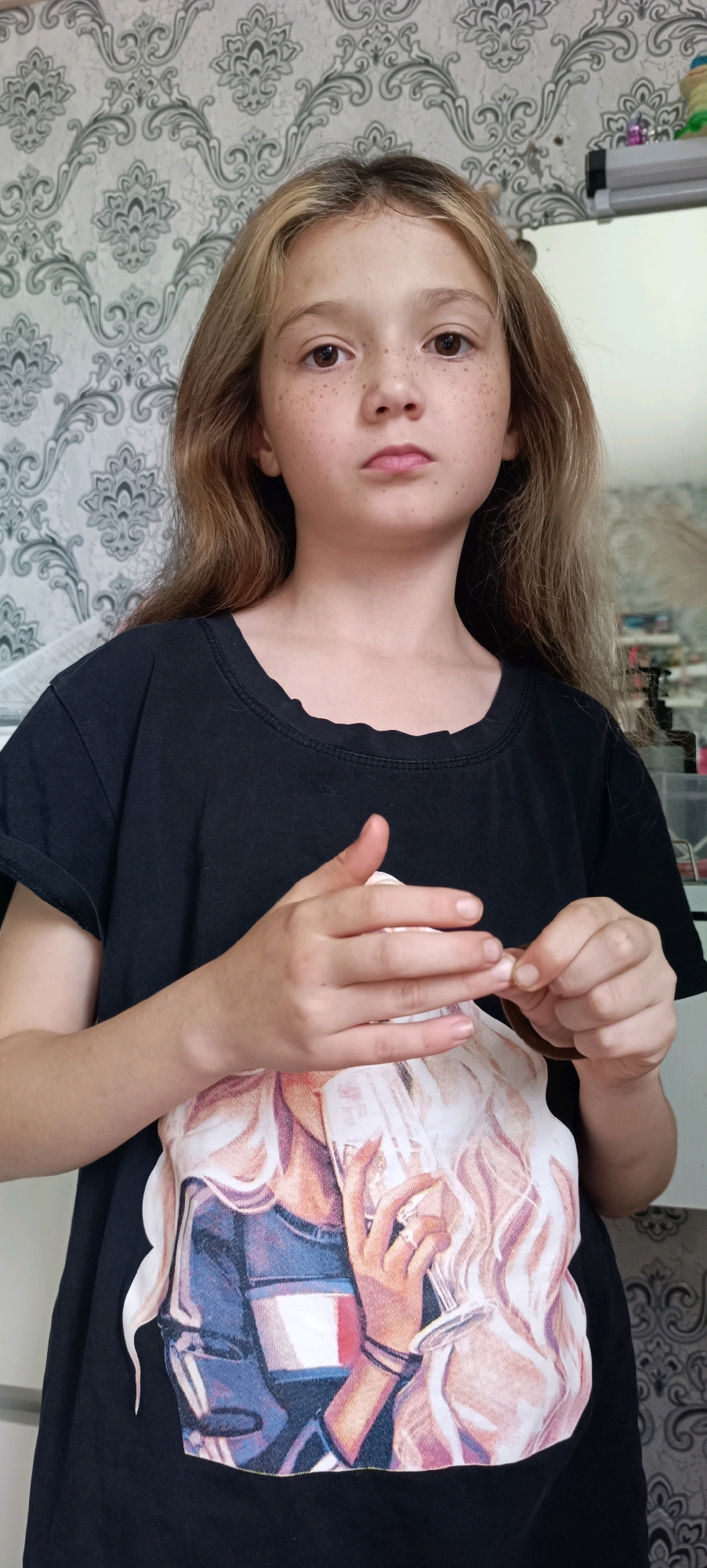 A young girl with blonde hair and freckles is shown from the chest up, looking directly at the camera. She is wearing a black t-shirt with a graphic print of a woman with blonde hair. The girl's hands are in the foreground, and she is fiddling with a brown hair tie. The background is a wall with a grey and white floral wallpaper pattern. The lighting appears to be natural, suggesting it is daytime. The scene is indoors, likely a home environment. The girl's expression is neutral.