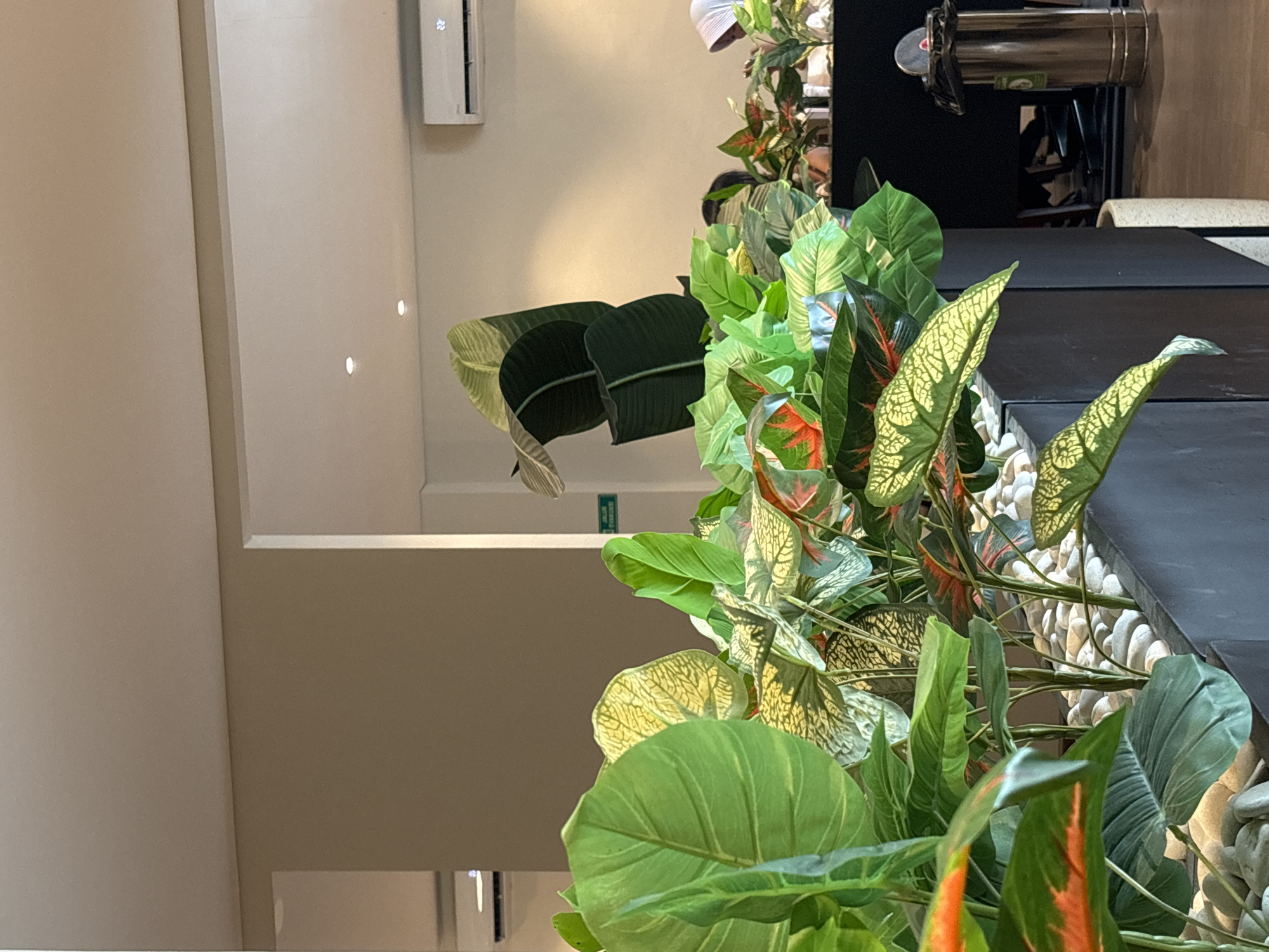 The image depicts an indoor scene with an abundance of artificial plants, primarily in various shades of green with some red and yellow accents. The plants are arranged in a planter box filled with white stones. In the background, a beige wall is visible, along with a white ceiling that has recessed lighting. To the right, a white air conditioning unit is mounted on the wall. Further to the right, a glimpse of a person wearing a light-colored headscarf and a dark garment is visible, suggesting the presence of other people. A tall, metallic trash can with a black plastic liner is also present in the mid-right portion of the image. The overall environment appears to be a clean, well-lit indoor space, possibly a cafe, office, or commercial establishment. The image does not provide any specific visual cues or landmarks that would allow for precise localization within West Jakarta, Indonesia.