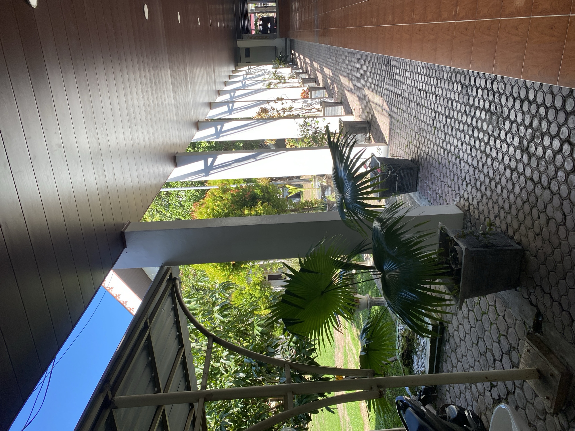 This is a ground-level, eye-level shot of a covered walkway or veranda in Banda Aceh, Indonesia. The walkway is lined with regularly spaced white columns casting sharp shadows on a patterned cobblestone floor. Along the walkway, numerous potted plants, primarily palm varieties, are arranged. To the left of the walkway, there is a covered parking area with a motorcycle visible. Beyond the parking area, lush green trees and grass are visible under a bright blue sky. The walkway appears to be part of a building with a brown, wood-paneled ceiling equipped with recessed lighting. The scene is well-lit by natural sunlight, creating a strong contrast between light and shadow.