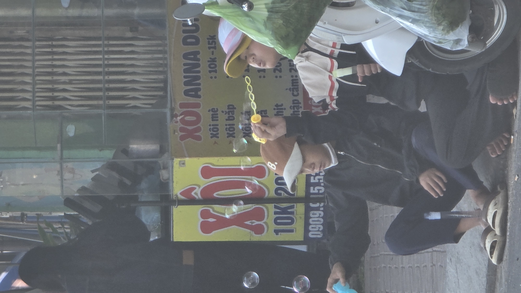 The image captures a street scene, likely in Bắc Nha Trang, Vietnam. Two individuals, possibly a father and child, are visible. The child, wearing a hat, is seated on a parked scooter. The other individual is blowing bubbles. In the background is a sign for "Xoi Anna Du," offering various types of xoi (sticky rice) for sale. The sign lists prices ranging from 10,000 to 25,000, possibly Vietnamese Dong. Bubbles float in the air. The individuals are barefoot, and wearing casual clothing.