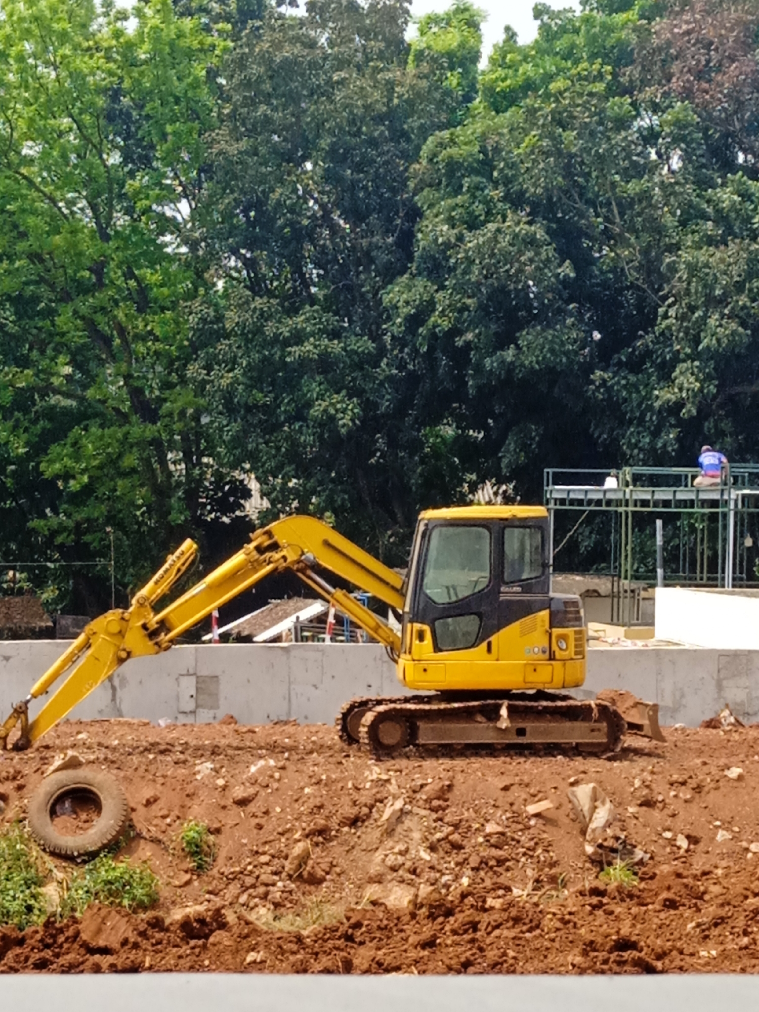 A yellow excavator is parked on a mound of red dirt. The excavator has a black cab with windows and a large yellow arm. A tire is lying on the ground in front of the excavator. In the background, there are trees and a construction site with scaffolding and workers. The scene is set in the city of Bekasi, Indonesia.