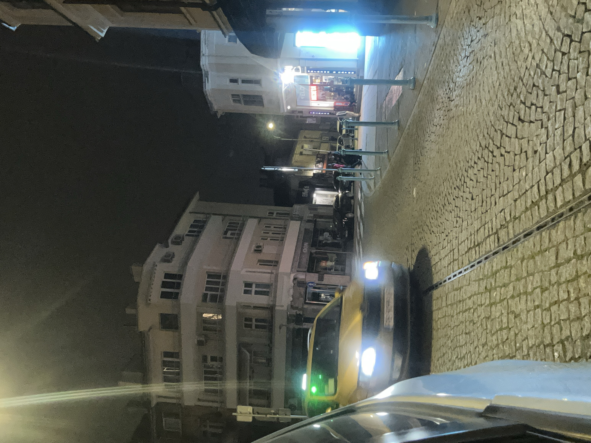 The image shows a nighttime street scene in Burgas, Bulgaria. The street is paved with cobblestones, and there are buildings on both sides. The building on the left is a multi-story white building with multiple windows and air conditioning units. On the right side of the image, a section of a building is visible with several windows and what appears to be a shop or storefront with bright lighting. There is a street light illuminating the area. There are parked cars along the street and what appears to be a tram line running along the street. A yellow car with its headlights on is positioned in the middle of the street. In the foreground, the hood of a silver-colored car is visible.