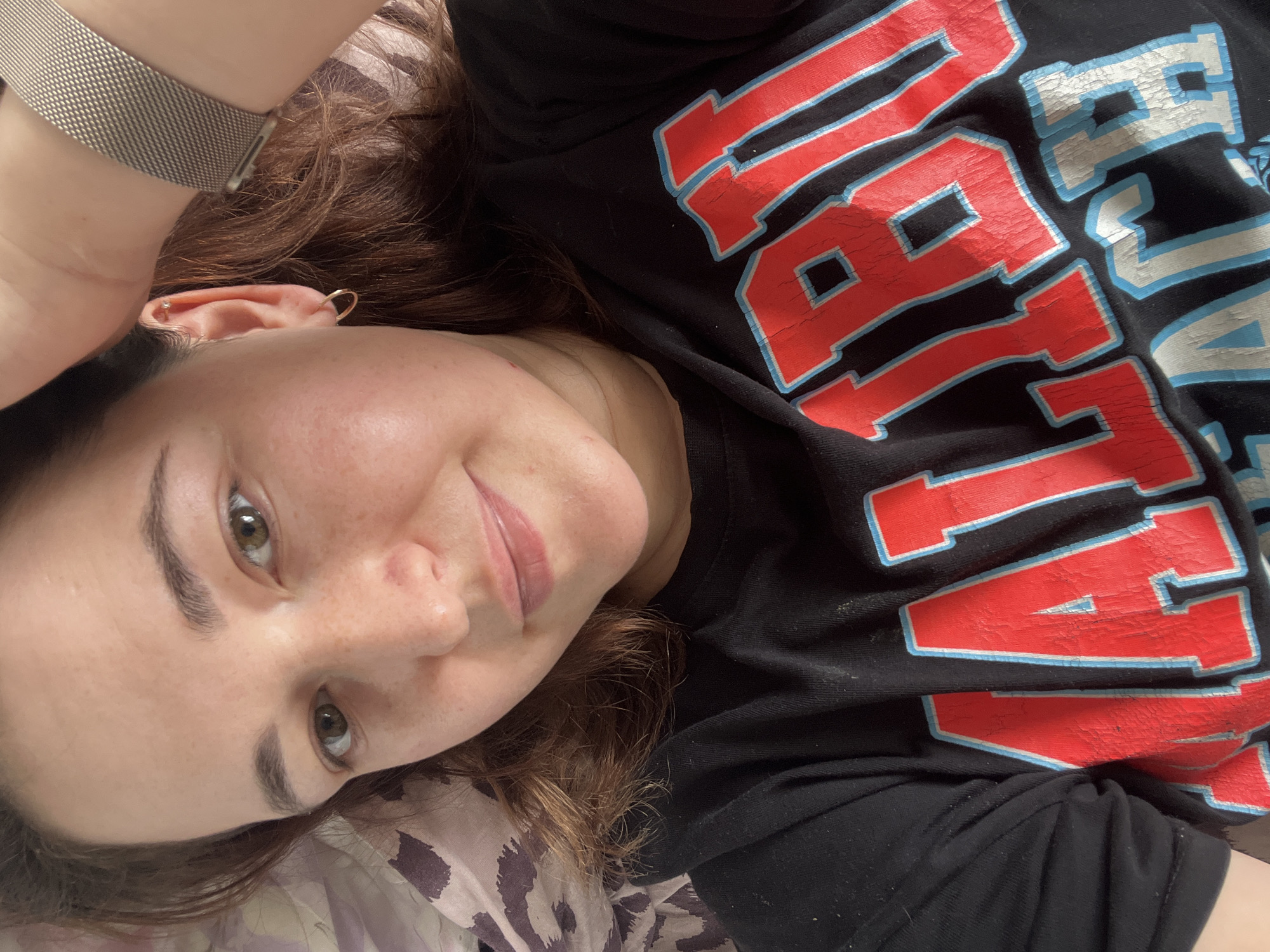 The media file is a close-up, angled selfie of a person lying down, likely indoors. The subject is a woman with brown hair, fair skin, and green eyes. She is wearing a black t-shirt with large, red and blue block lettering that appears to say "NAVY". There is a metallic watch on her left wrist and earrings in her ears. The background is blurred, but it appears to be a patterned surface, possibly bedding. The lighting suggests it is daytime, and the overall mood is relaxed and casual. There are no obvious location cues to specifically place this in Chelyabinsk, Russia, beyond the provided context.