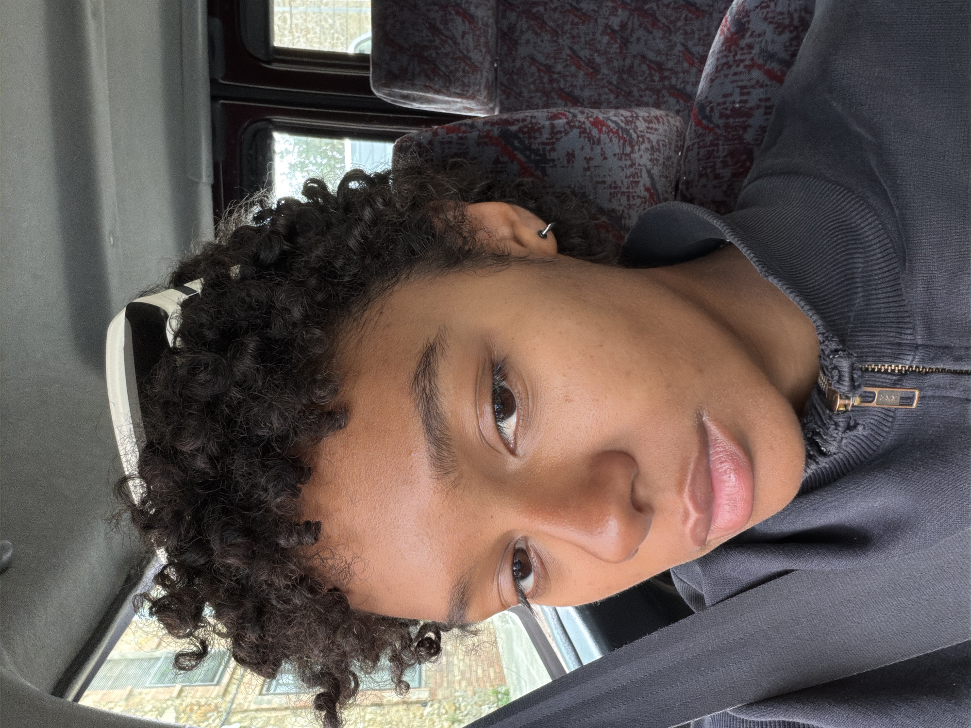 A close-up, eye-level, indoor portrait shows a person with brown skin and curly black hair, wearing a dark-colored zip-up hooded jacket and a seatbelt. The person has brown eyes and a small hoop earring in their left ear. The background shows a bus or van interior with window frames and upholstered seats. The location is possibly Pals, Spain. The person is looking directly at the camera.