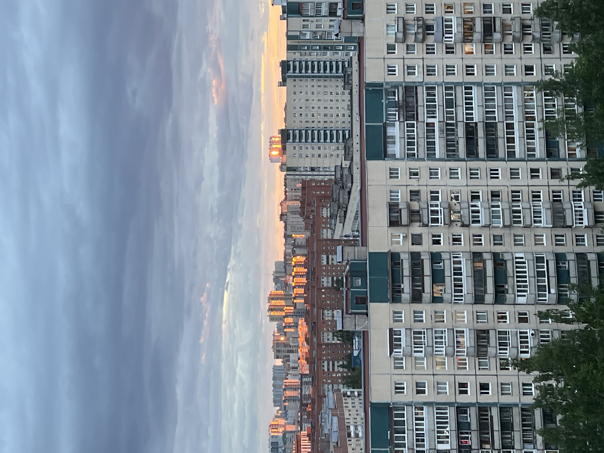 The image shows an urban landscape with a cityscape. On the right, a tall residential building with many windows is visible. Some windows are open. To the left of this building, a series of other buildings stretch into the distance, some reflecting the orange light of the setting sun. The sky is cloudy, with hues of gray and blue, mixed with orange and pink towards the horizon, where the sun appears to be setting. The overall impression is that of an evening scene in a densely populated urban area. Saint Petersburg, Russia.