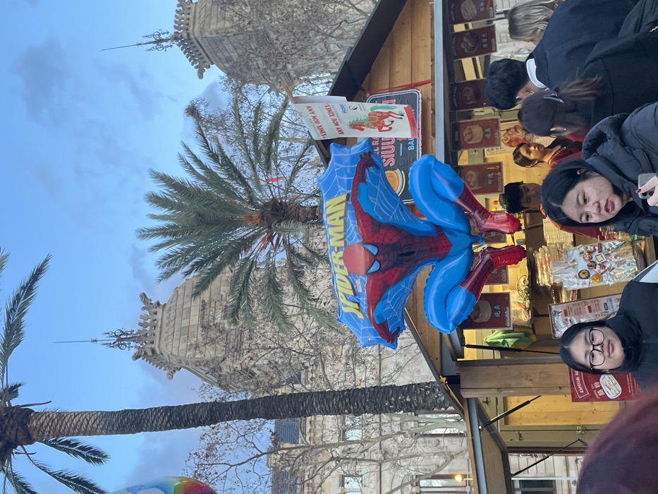 An outdoor scene depicts a building with spires, tall palm trees, and a vendor stall. A Spider-Man balloon prominently hangs from the stall. People, including two girls, are browsing or standing near the stall. A sign or poster with text and images is affixed to the stall's structure. The overall lighting suggests it is daytime, possibly dusk.